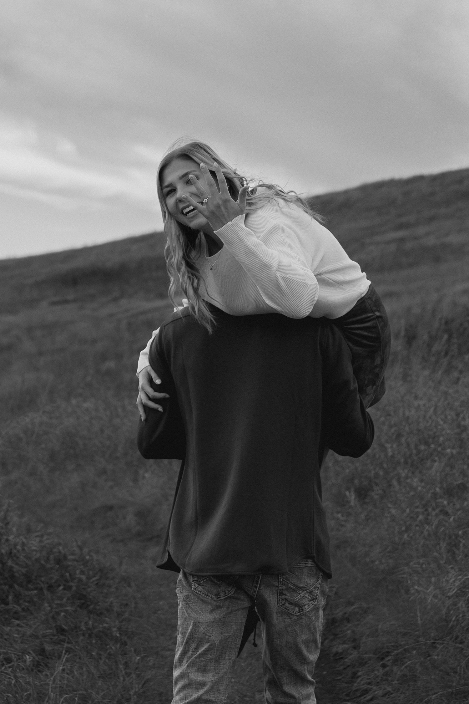 man picking up girl and taking her away on walking path as she hangs over shoulder and laughs while holding up her engagement ring to show