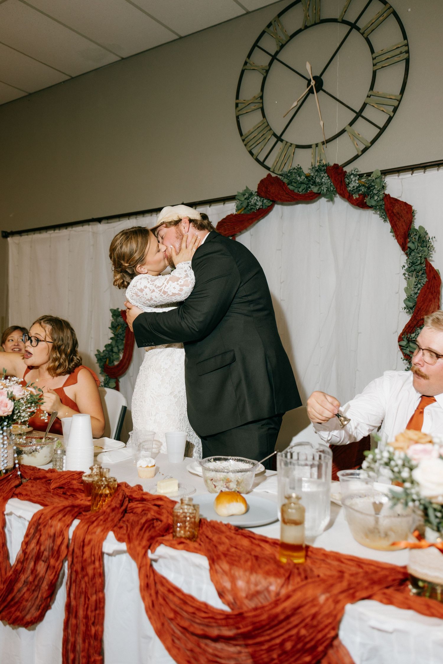 Bride and groom share a kiss at wedding reception table decorated with red fabric and greenery arch.