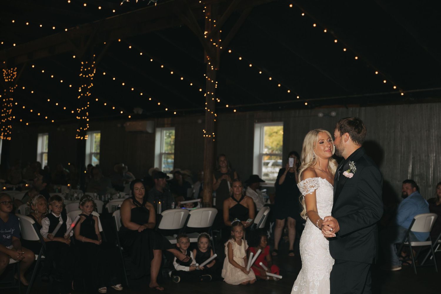 Bride and groom share first dance at elegant indoor wedding reception with string lights and seated guests.