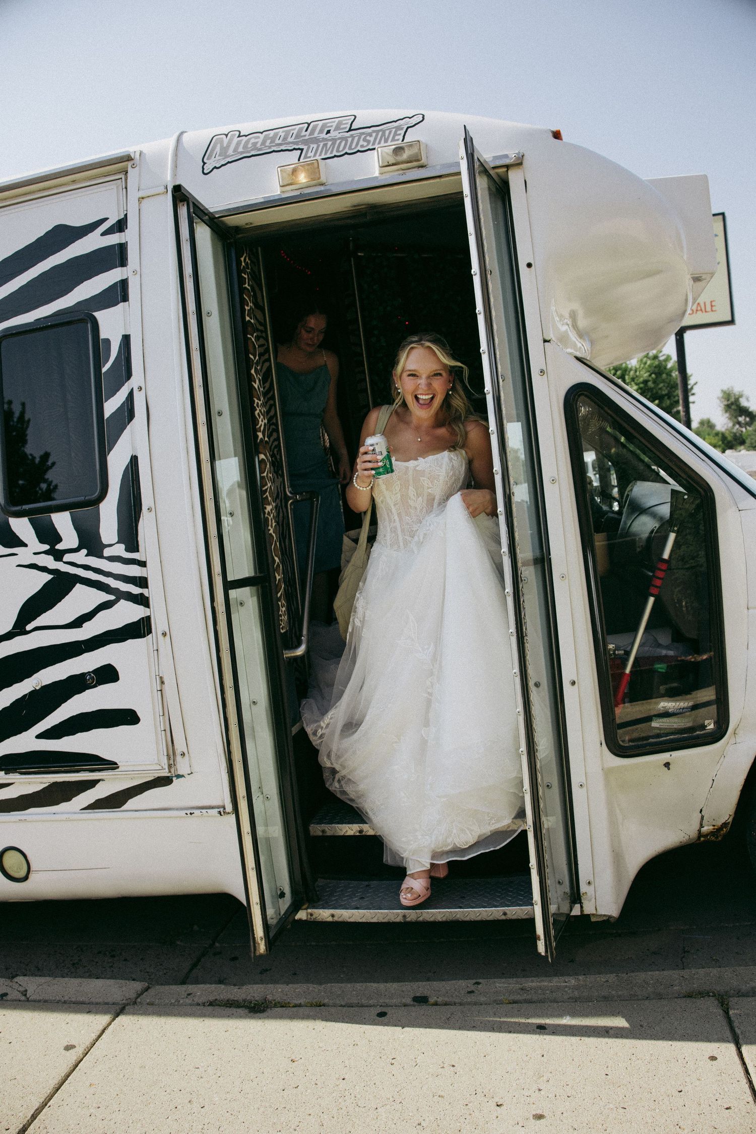 Excited bride in white gown stepping off zebra-print party bus holding a drink on her wedding day.