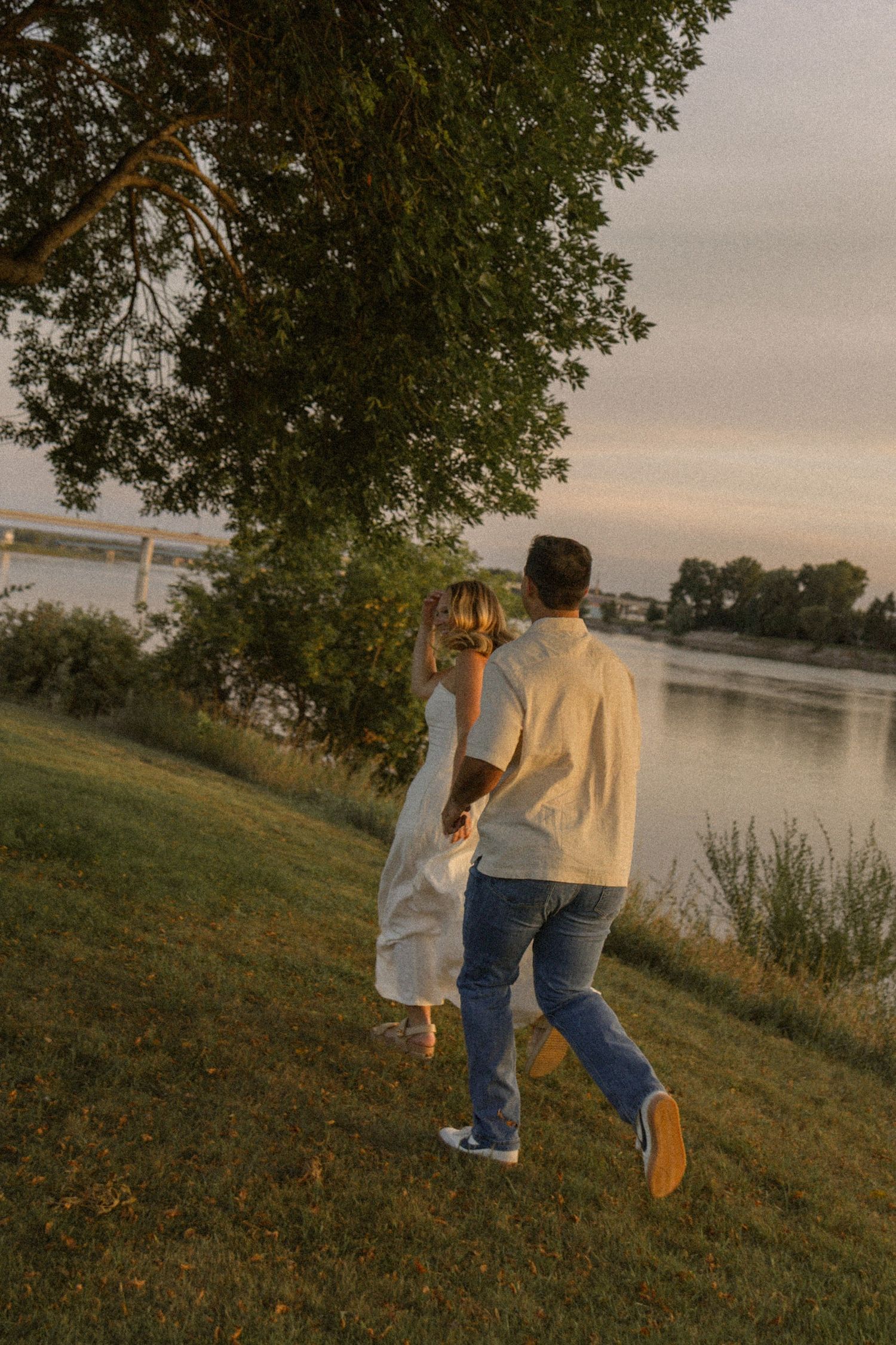 A couple walks hand in hand along a lakeside path during golden hour.