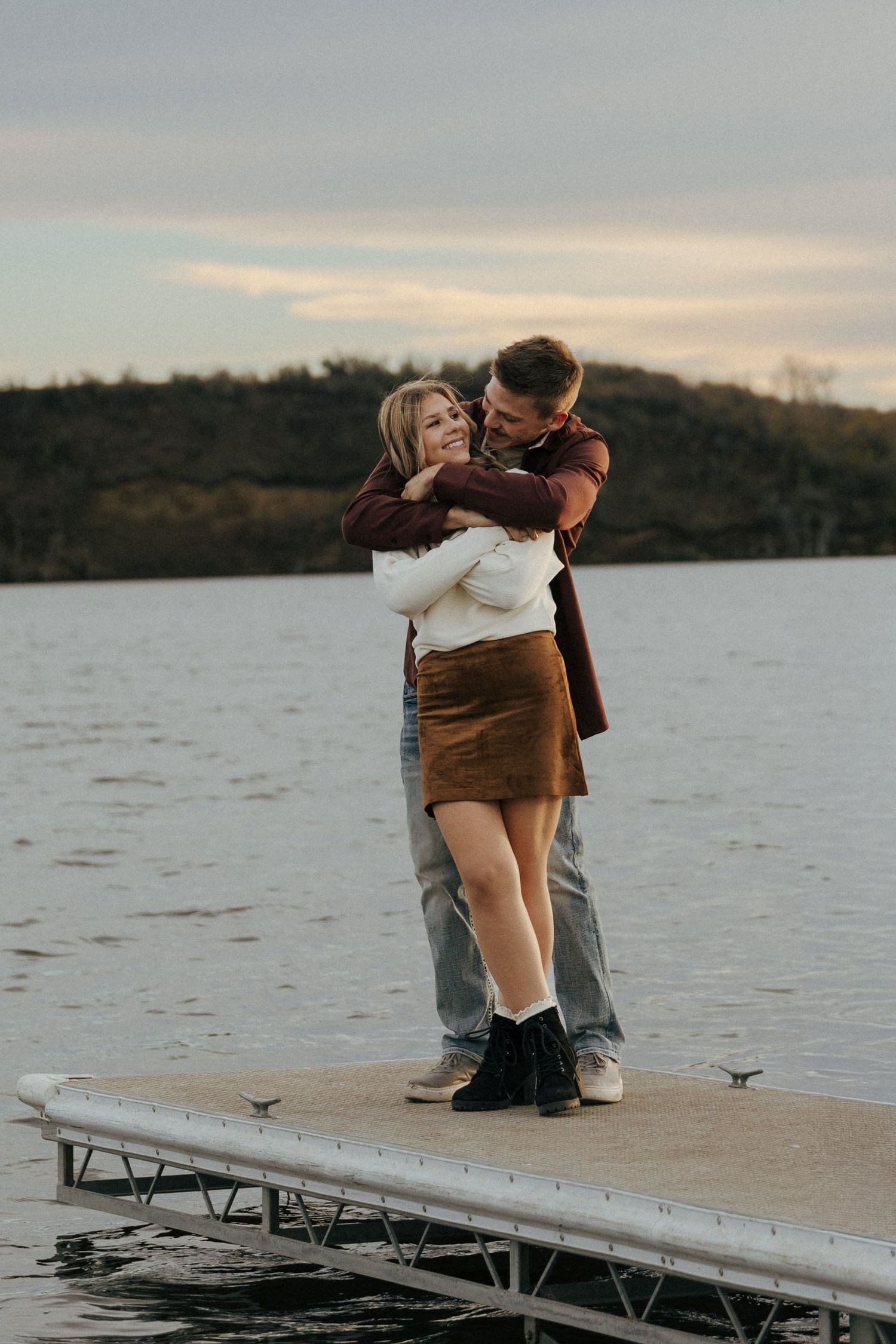 A couple shares an intimate moment on a wooden dock by a lake at sunset.