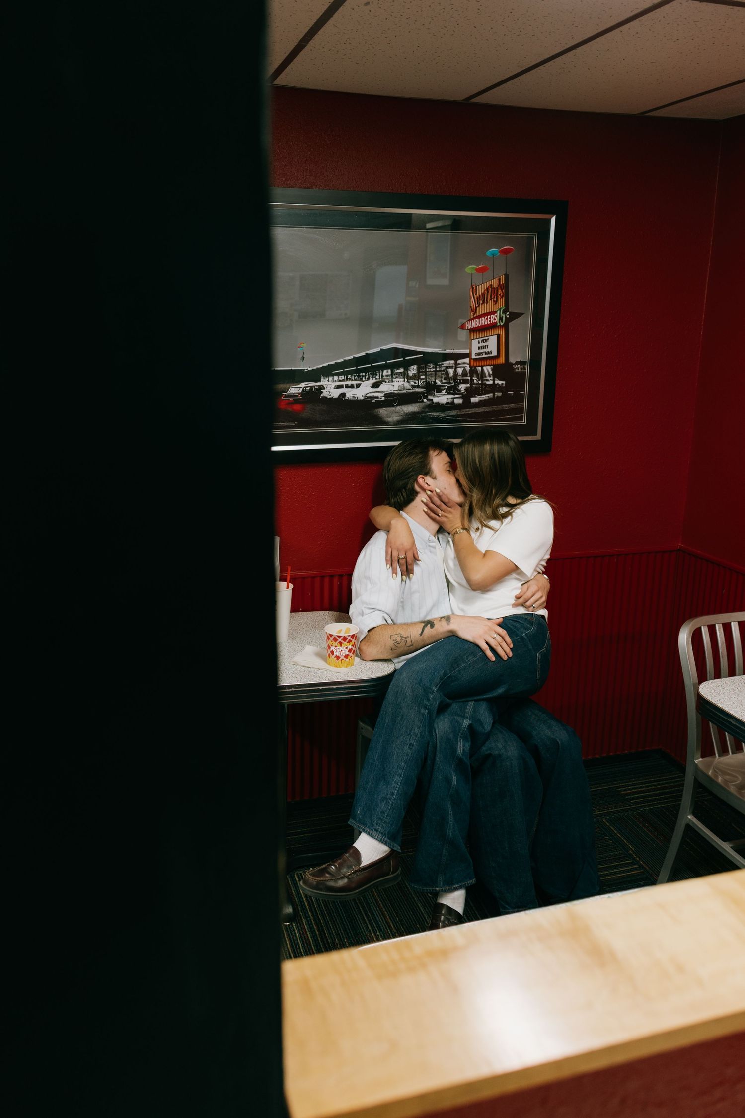 Couple sharing a kiss in a red diner booth, photographed through a doorway in warm candid style.