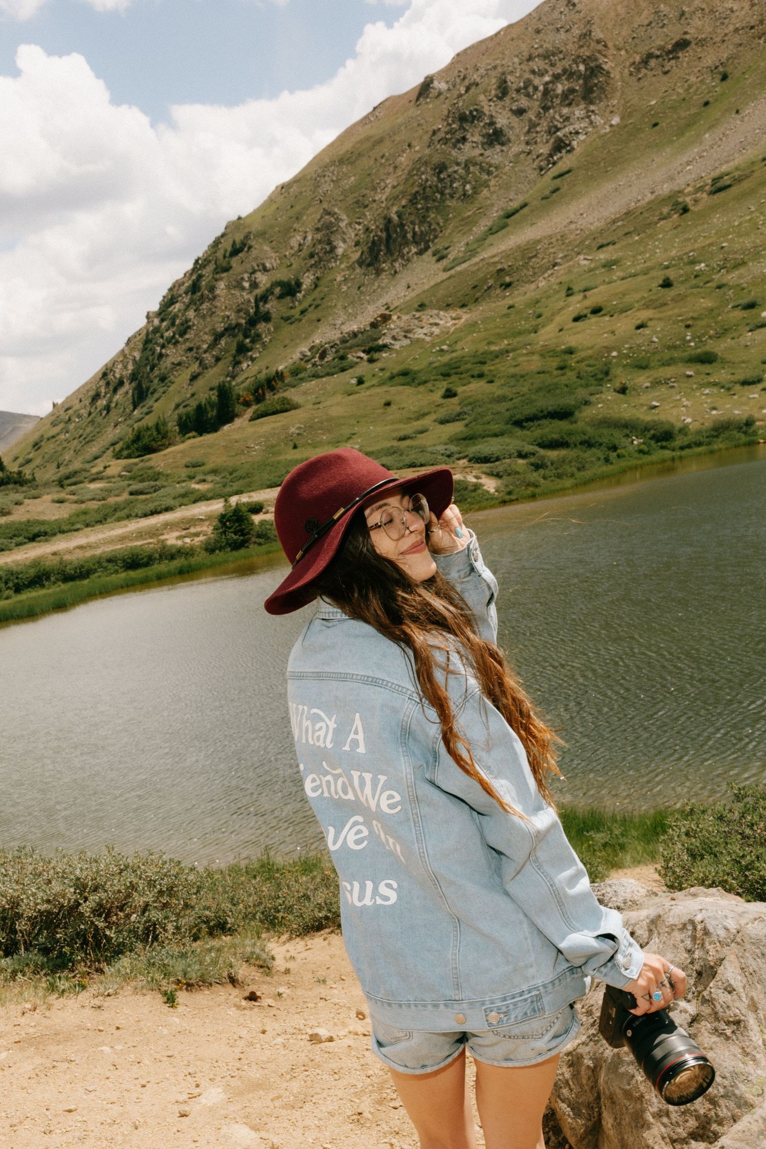 Woman in red hat and denim jacket holds camera by mountain lake.