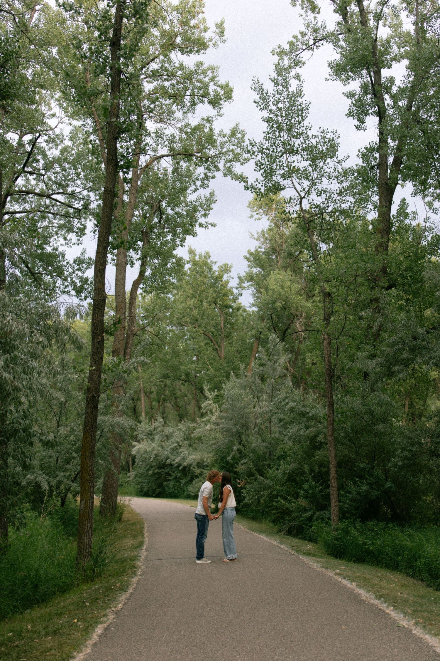 Couple holding hands on a tree-lined park path surrounded by lush green foliage under a cloudy sky.