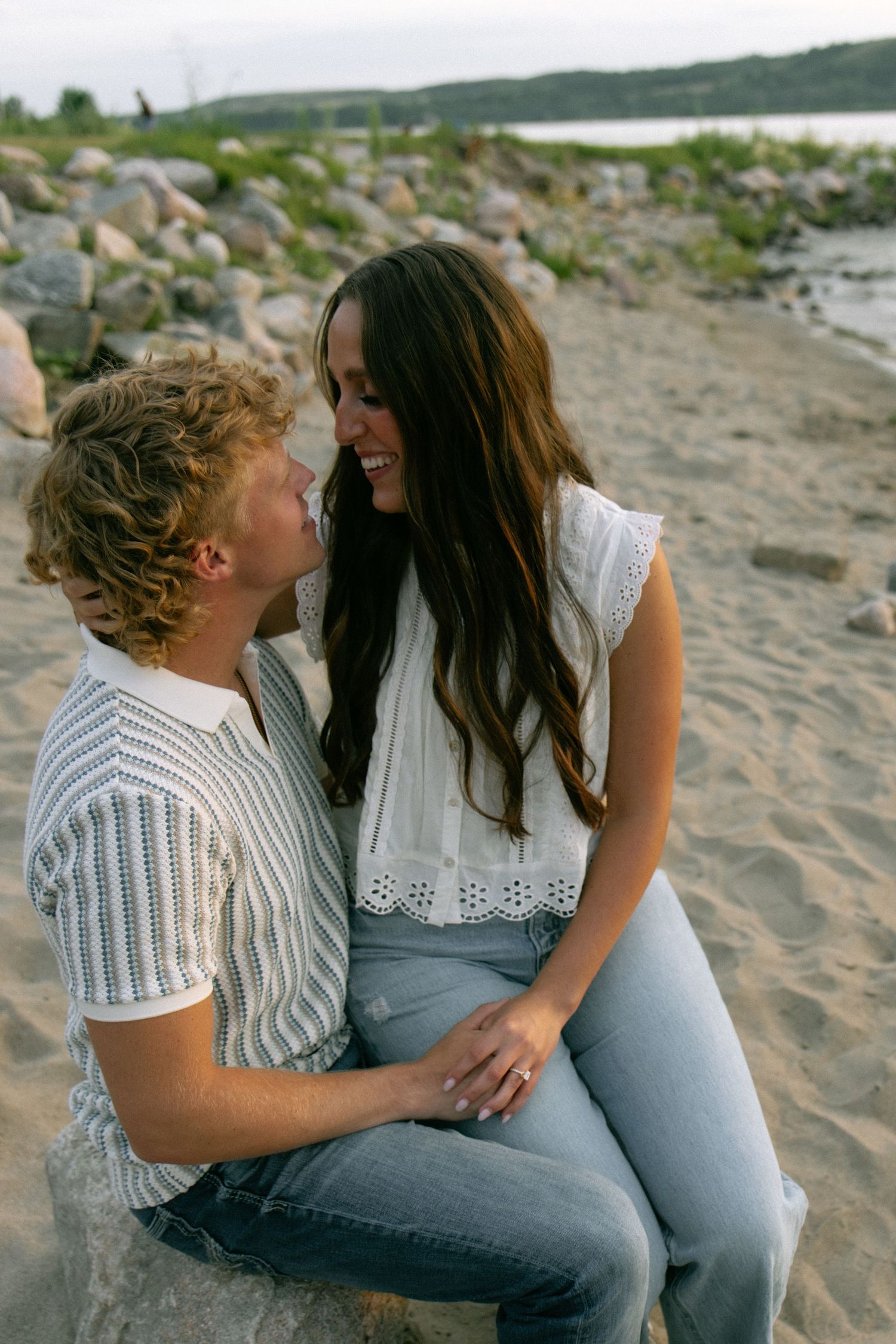 A couple shares a romantic moment on a sandy beach, smiling at each other while sitting among rocks near the water.