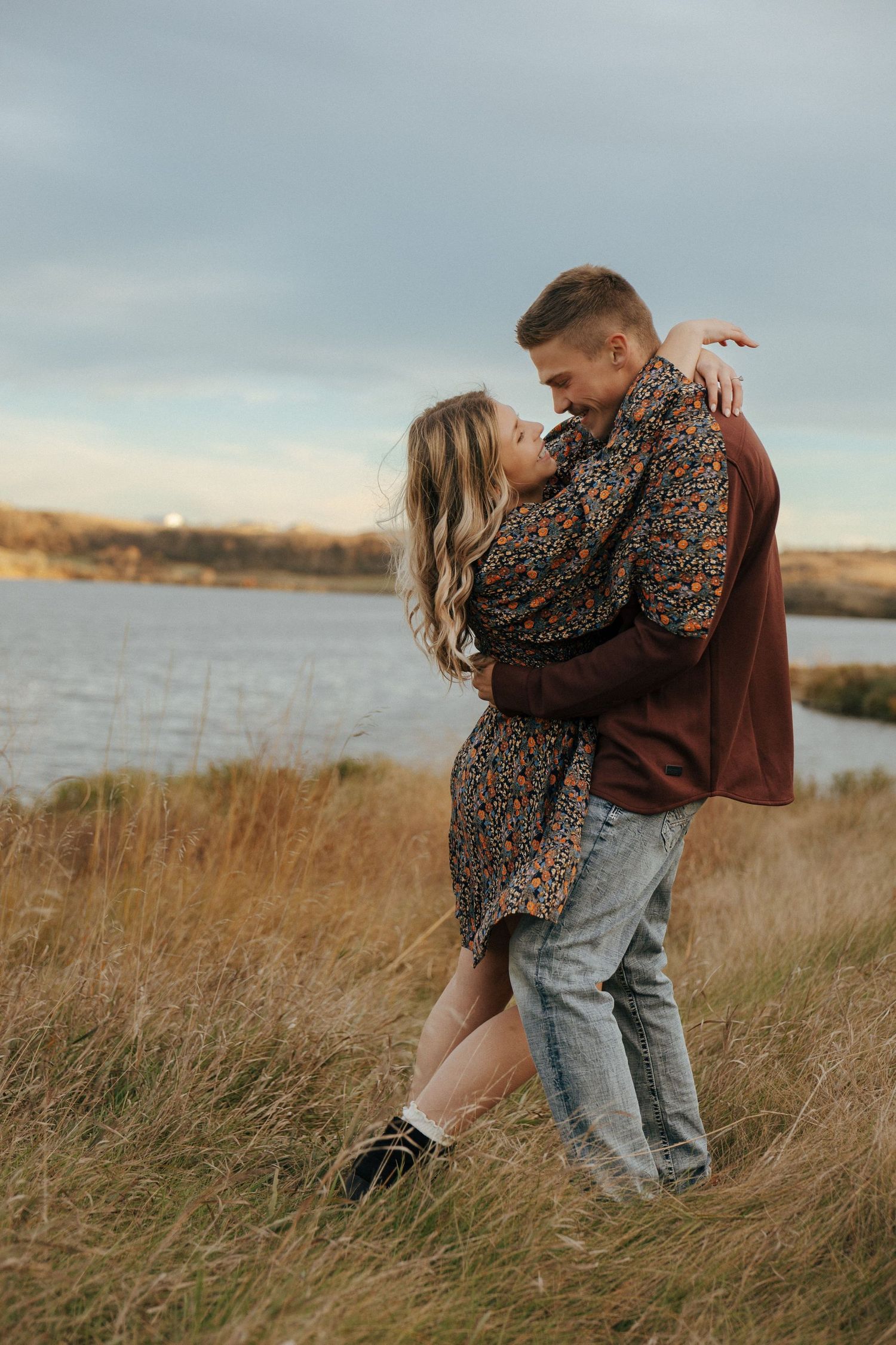 A romantic couple embraces in a field of tall grass at sunset near a lake.