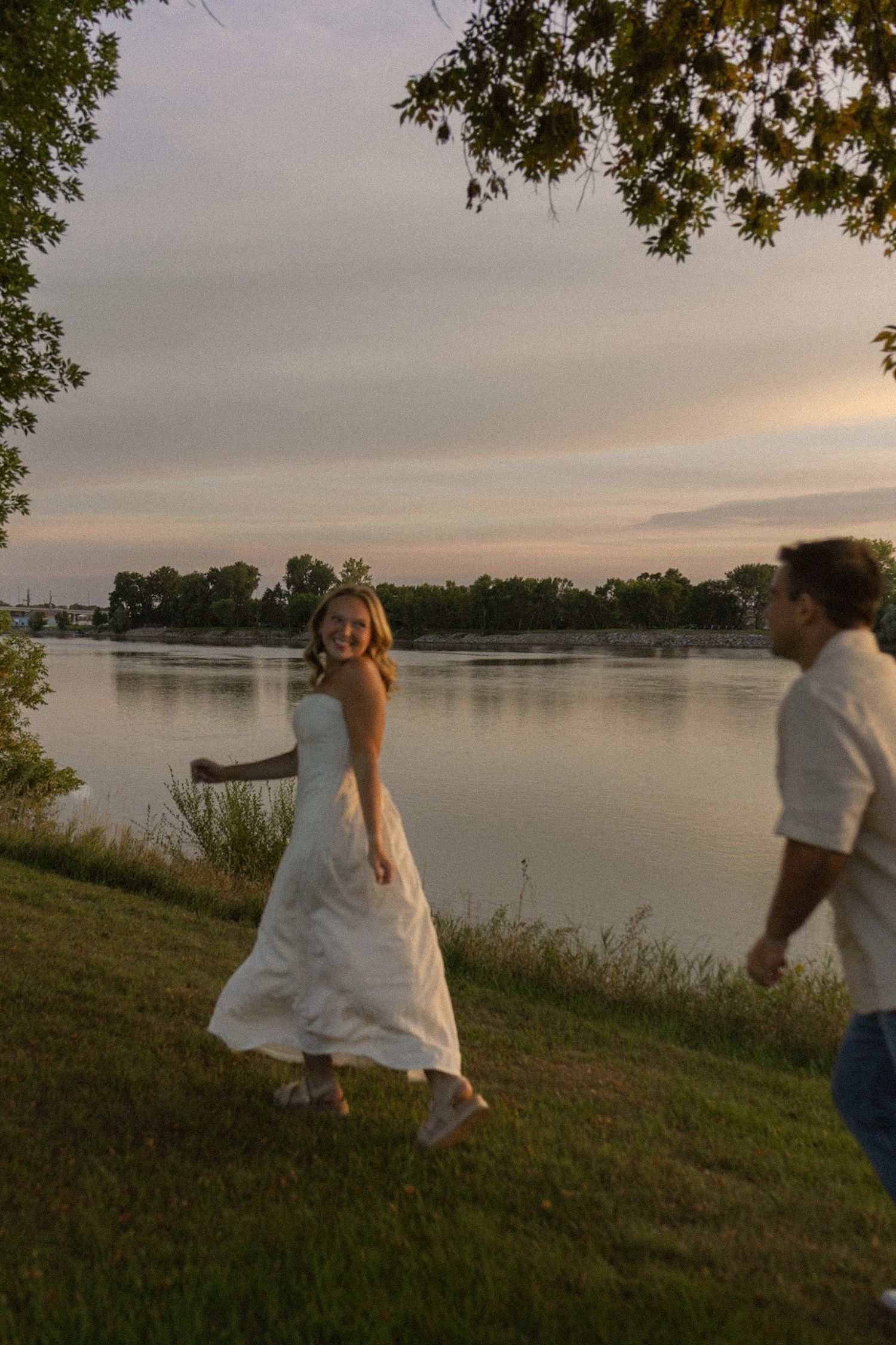 A couple plays and dances on a grassy slope near a lake during sunset.