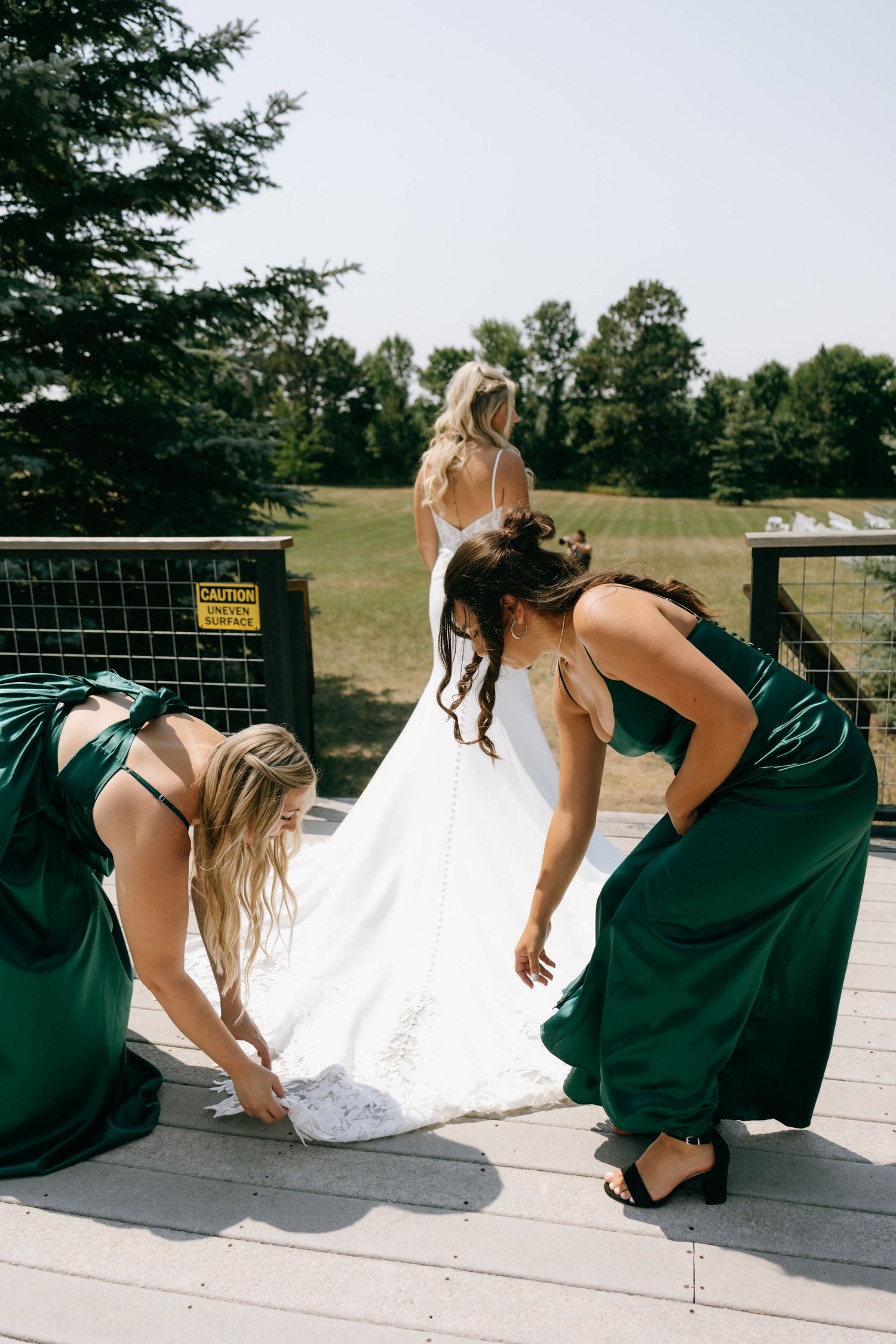 Two bridesmaids in emerald green dresses adjust a bride's white wedding gown train on an outdoor deck.