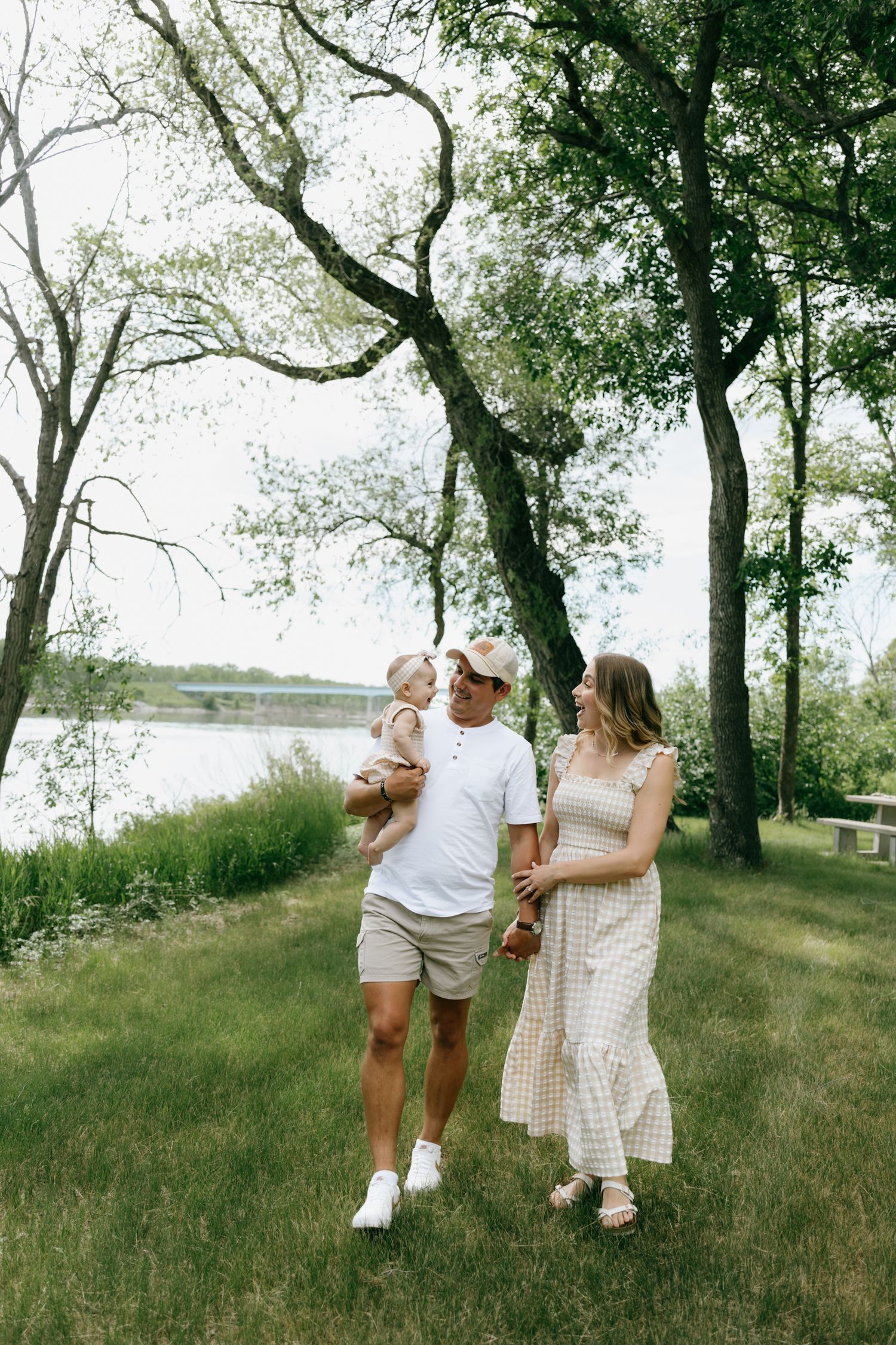 A young family with a baby walks outdoors near a lake surrounded by lush green trees on a sunny day.