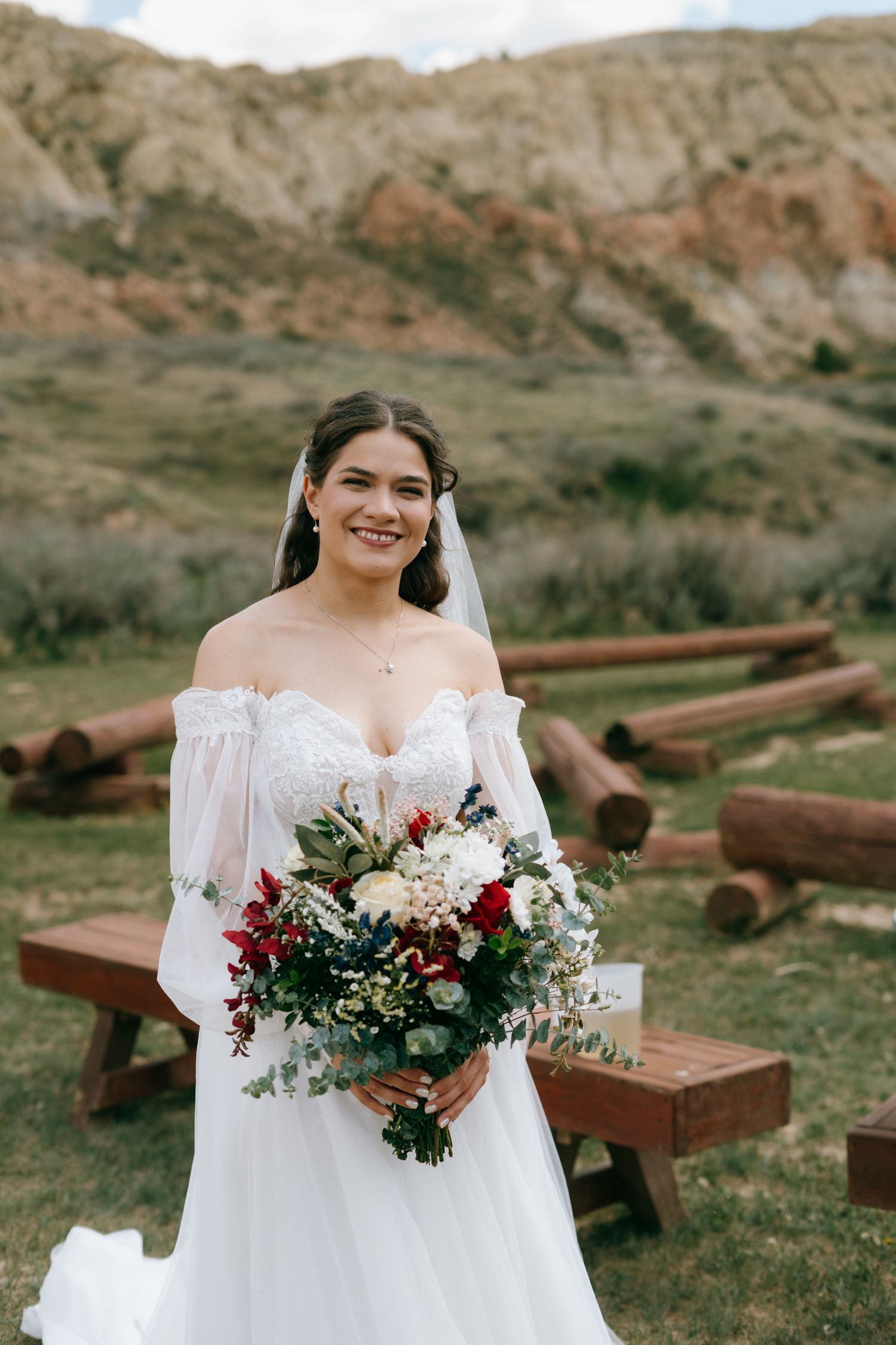 Smiling bride in off-shoulder lace gown holds red and white bouquet outdoors near rustic log benches and rocky hills.