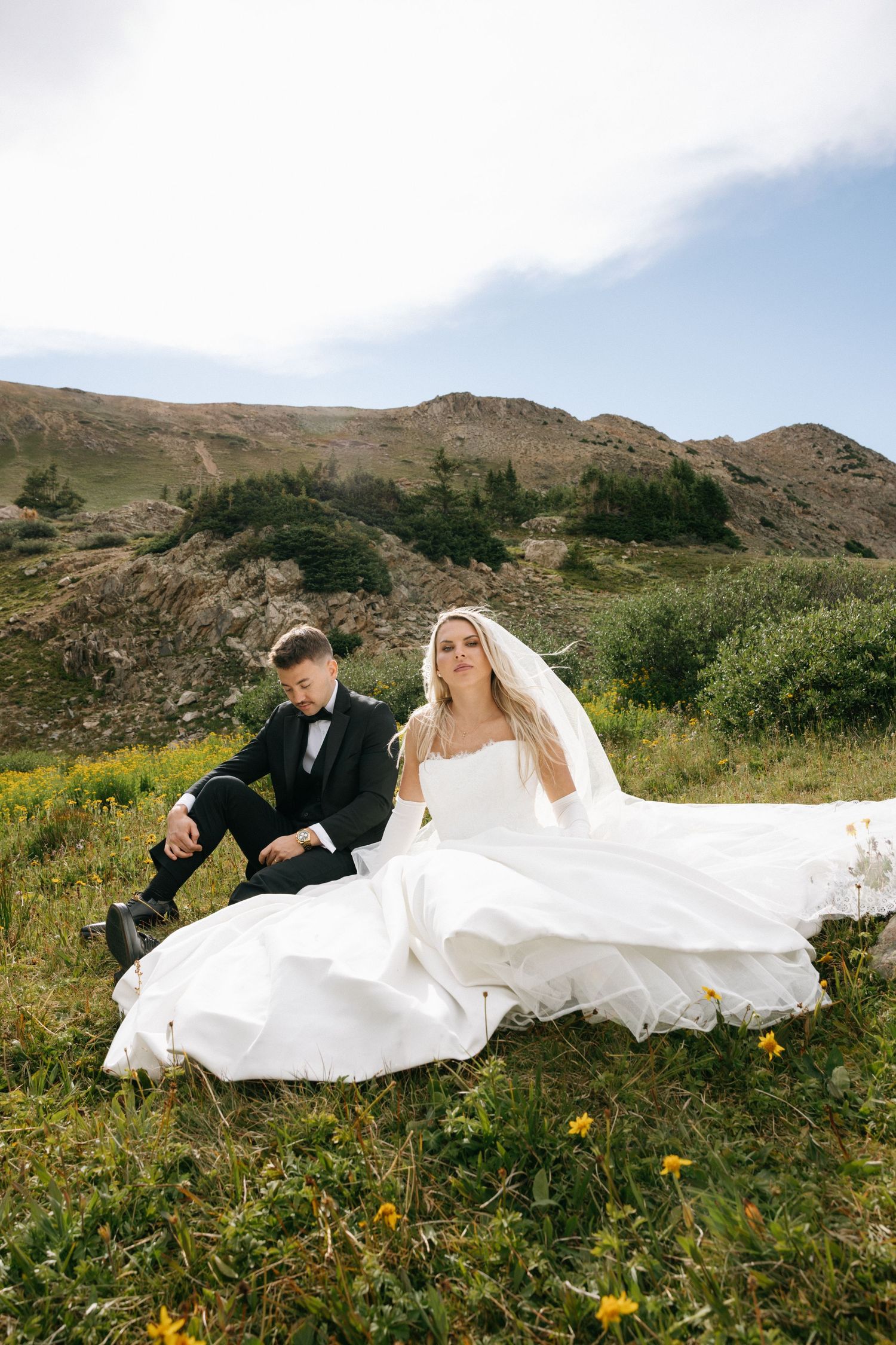 Bride in white gown and groom in black tuxedo sitting on green hillside with mountains backdrop.
