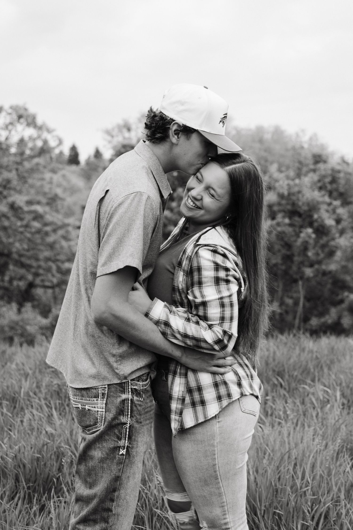 Black and white photo of a couple embracing and smiling in a field, man kissing womans forehead outdoors.