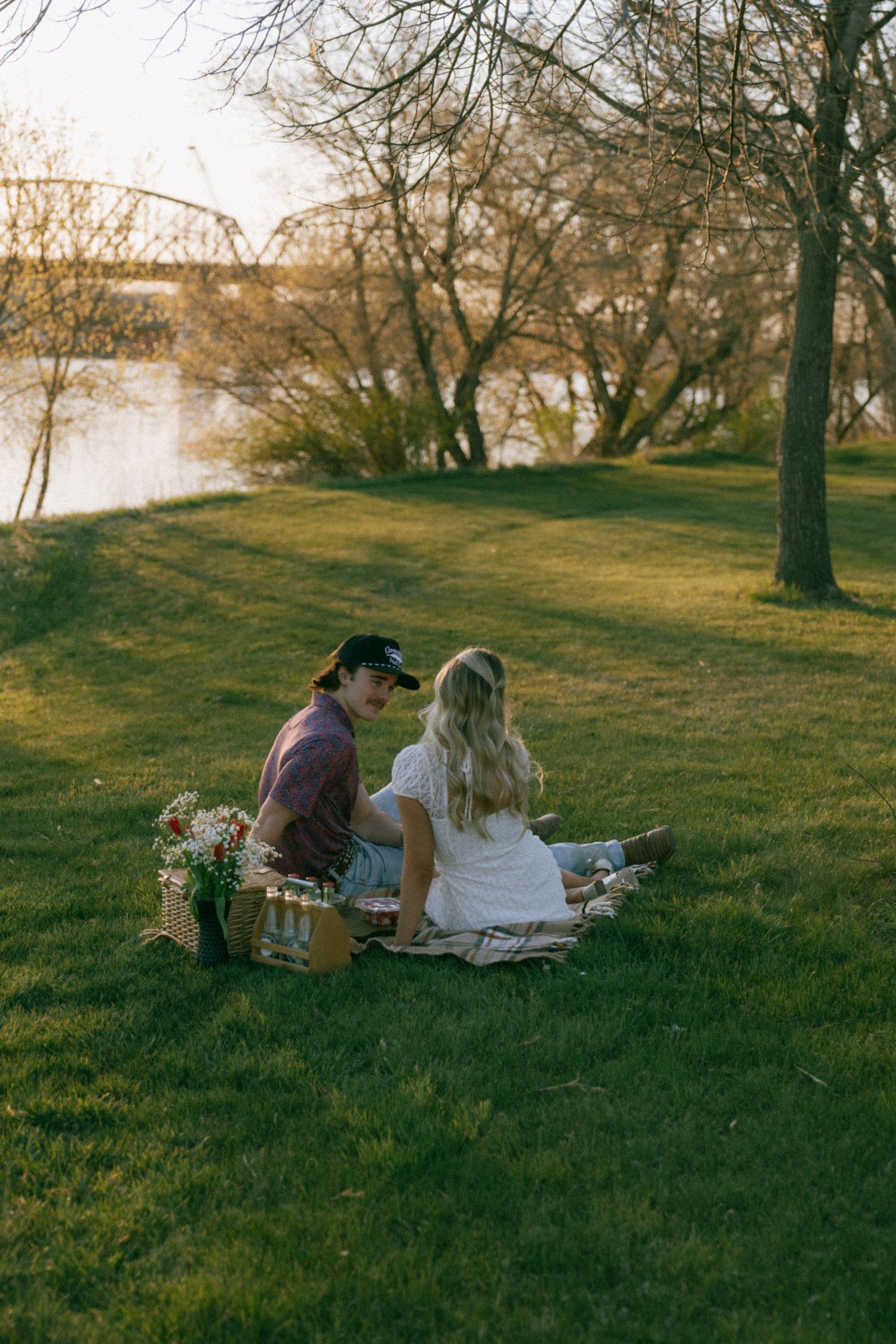 Couple sitting on a blanket having a picnic on green grass near a river at sunset.