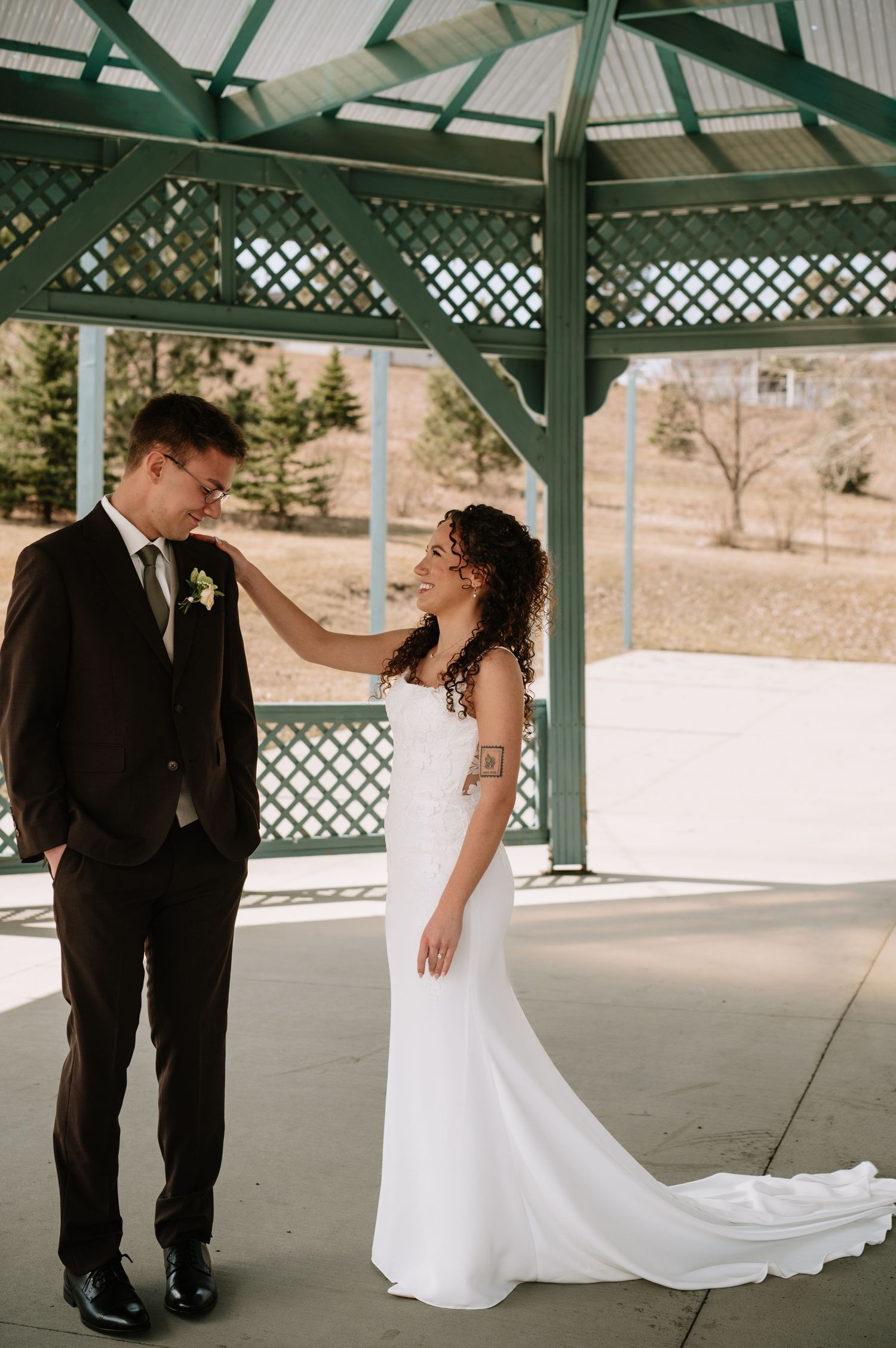 Bride in white gown touches groom's shoulder under green gazebo on their wedding day.