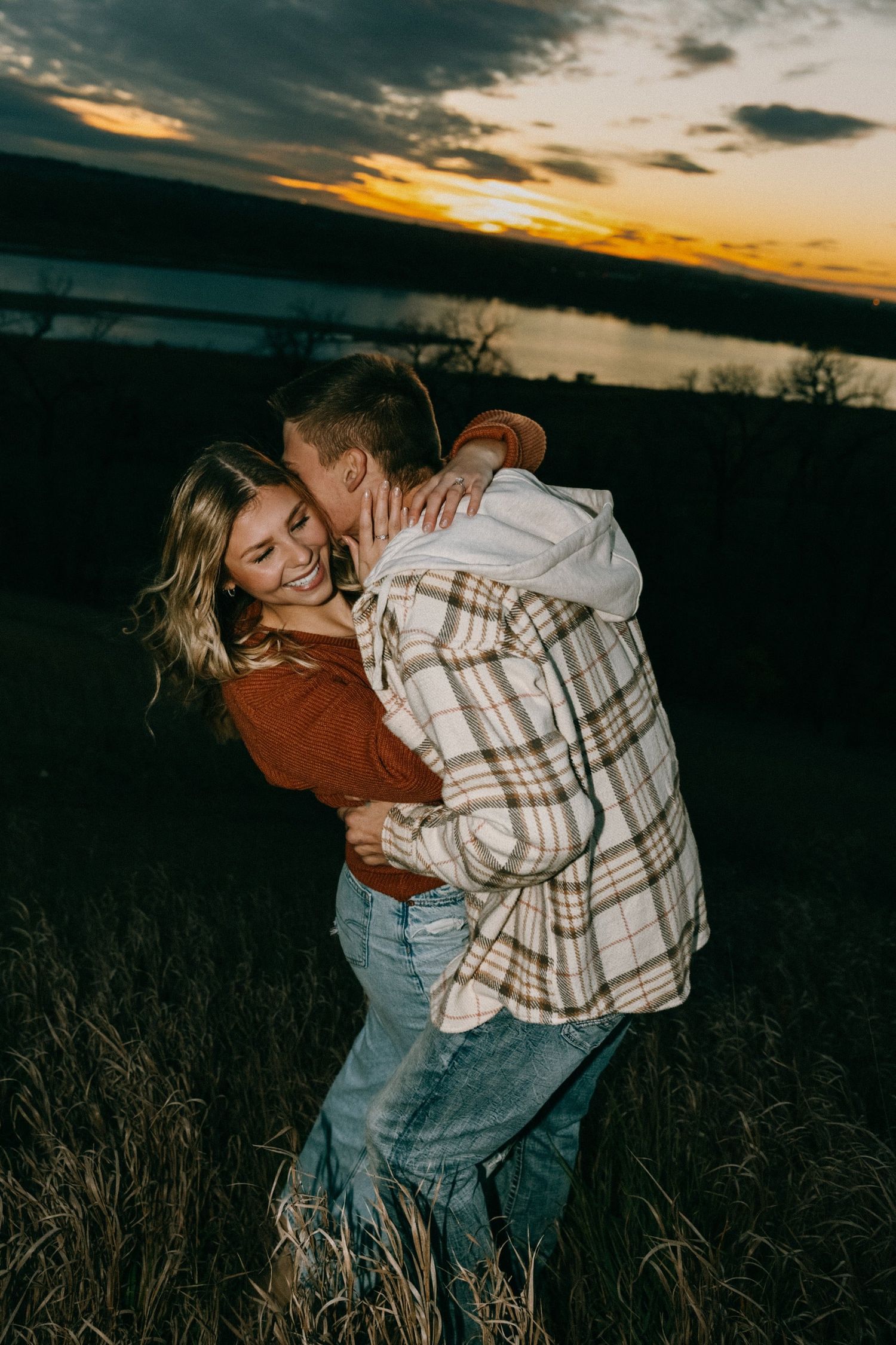 Couple in rustic attire share a romantic dance in tall grass at sunset.