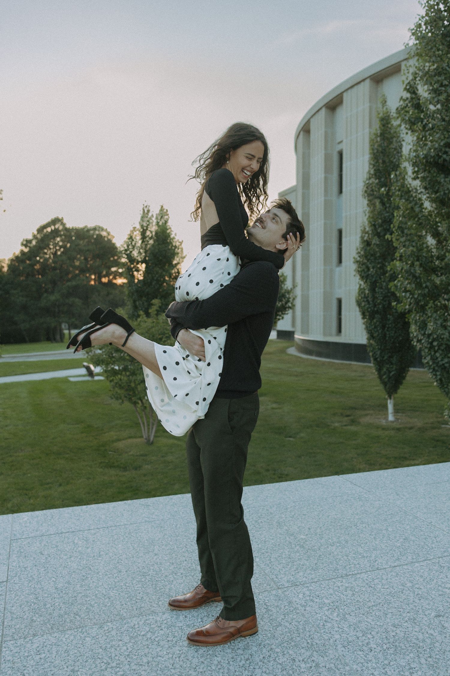 A couple in formal attire embraces and spins on a light colored surface at sunset.