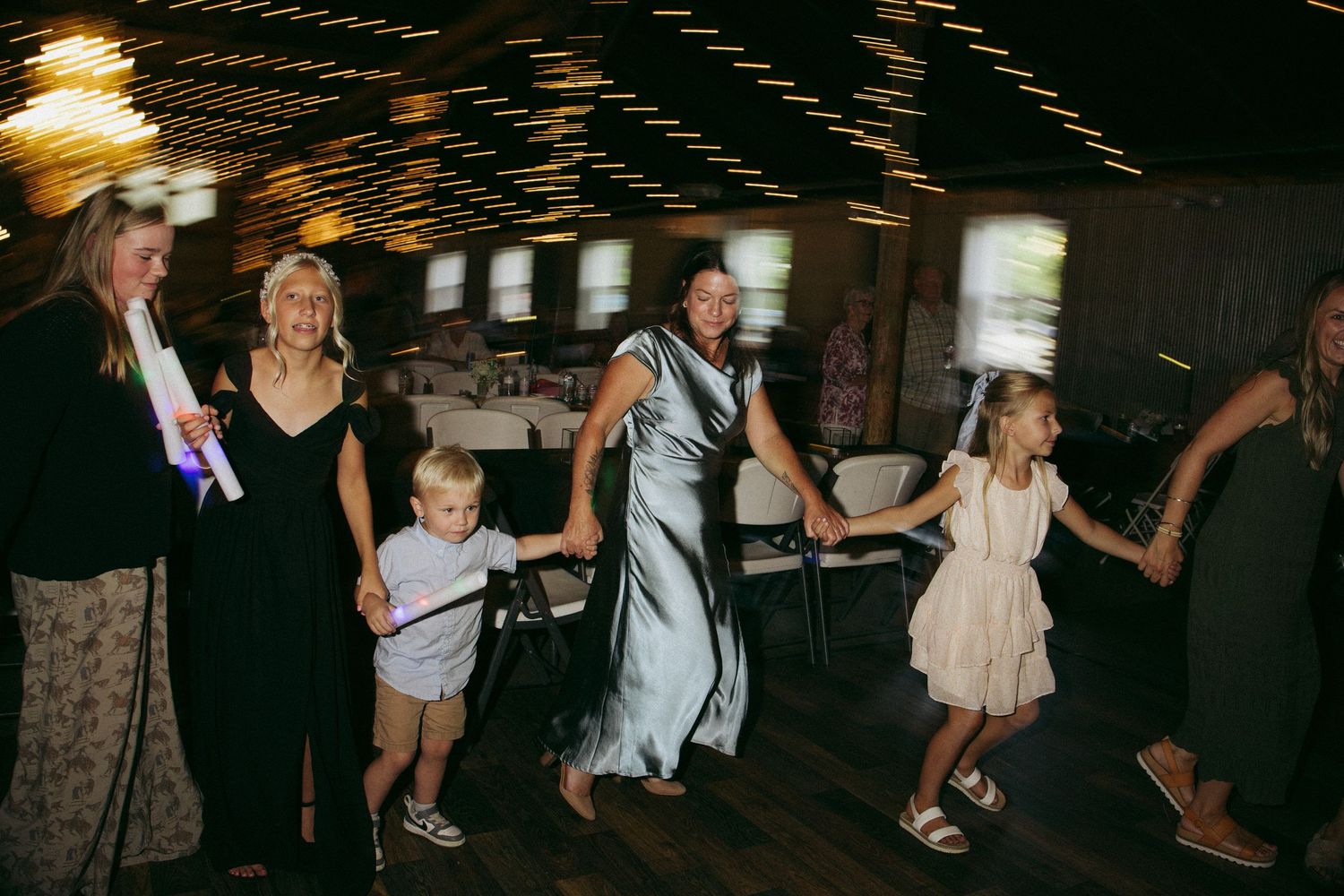 Wedding guests and children dance together joyfully inside rustic reception venue.