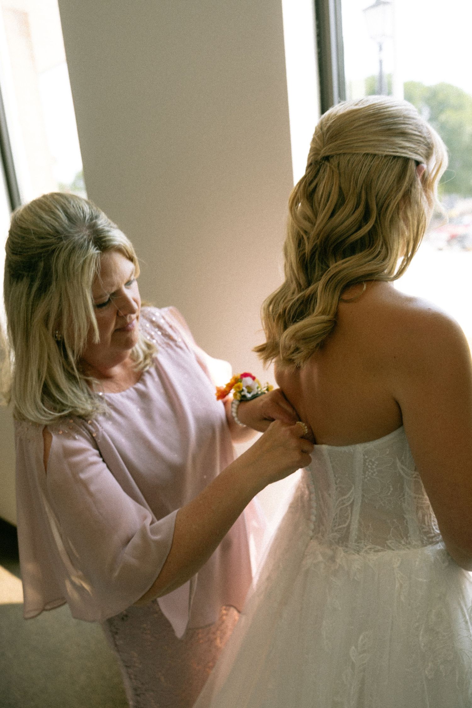 Mother helping bride button up her lace wedding dress near a bright window on the wedding day.