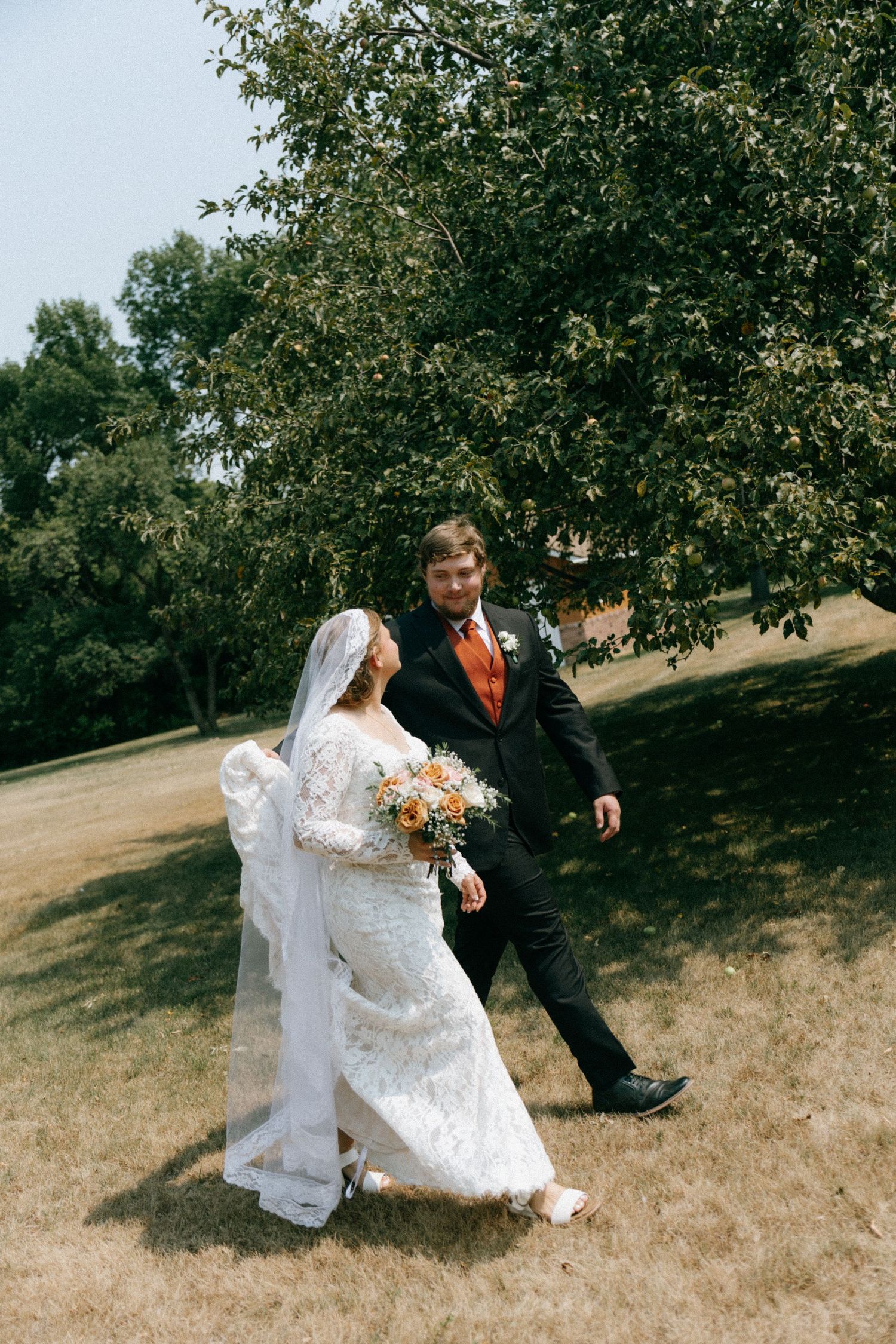 Bride and groom walking outdoors on grass near trees, bride in lace dress with veil, groom in black suit with orange tie.