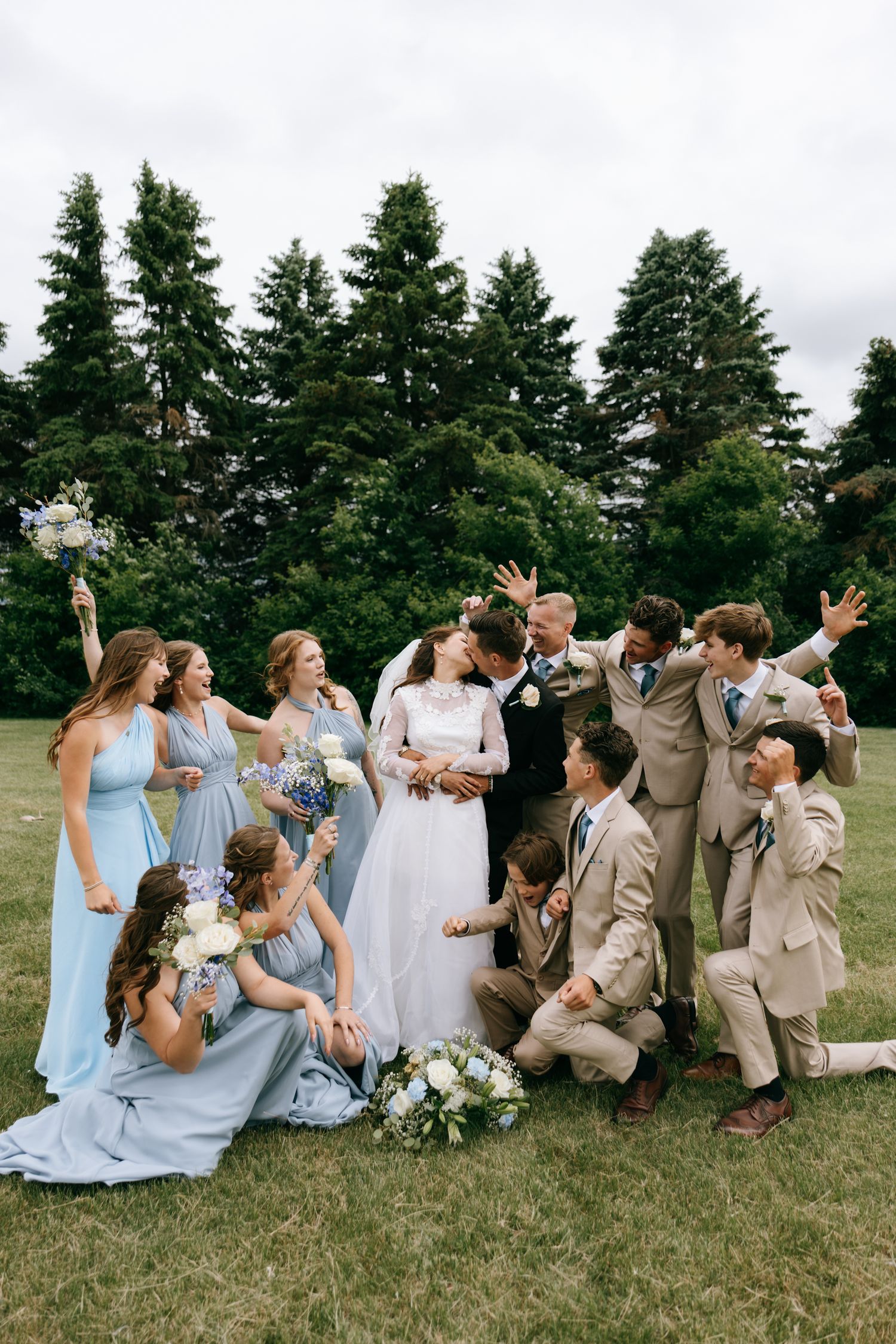 Wedding party celebrates bride and groom kissing outdoors, bridesmaids in blue dresses and groomsmen in tan suits on green lawn.