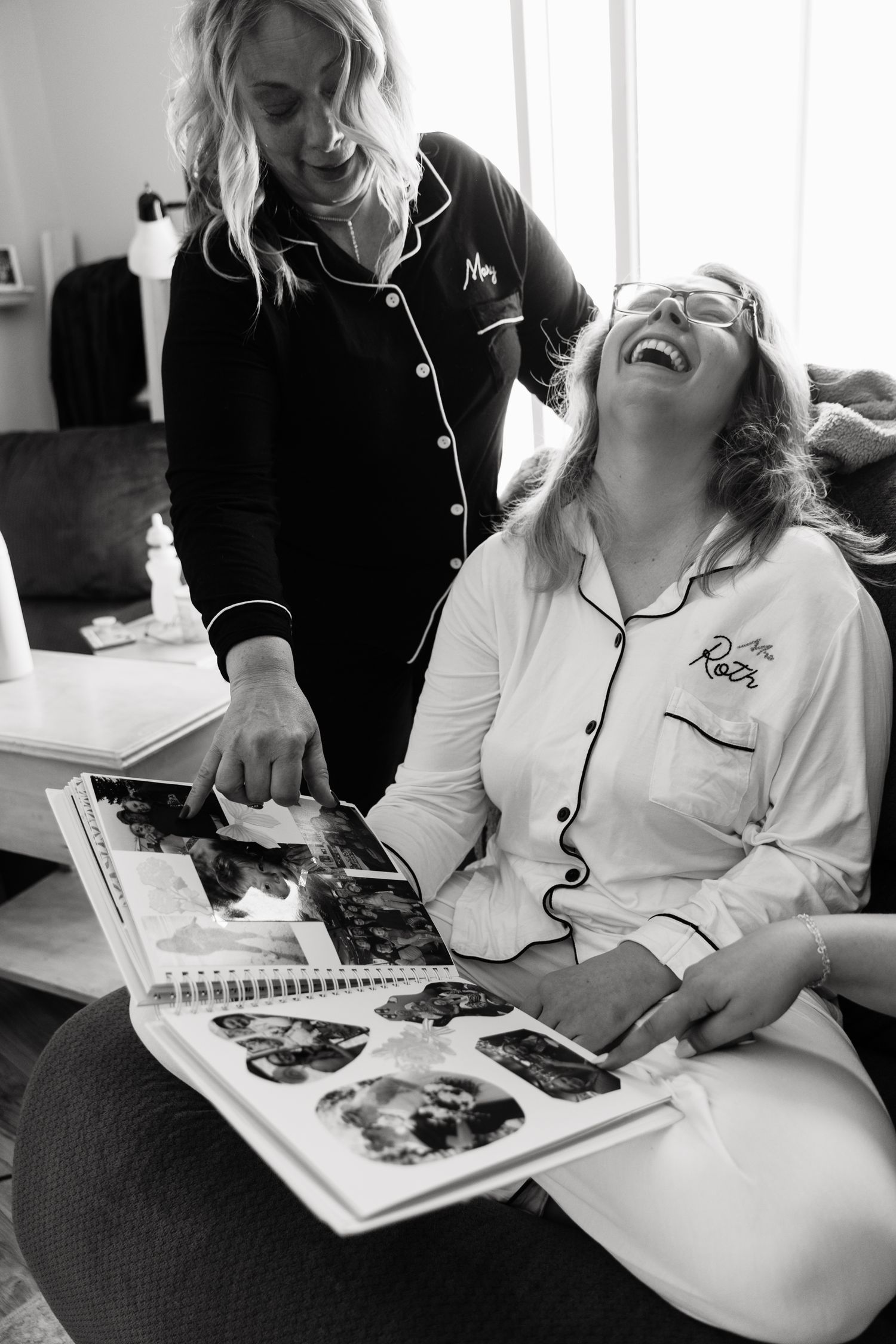 Two women in pajamas laughing while looking at a photo album together in black and white.
