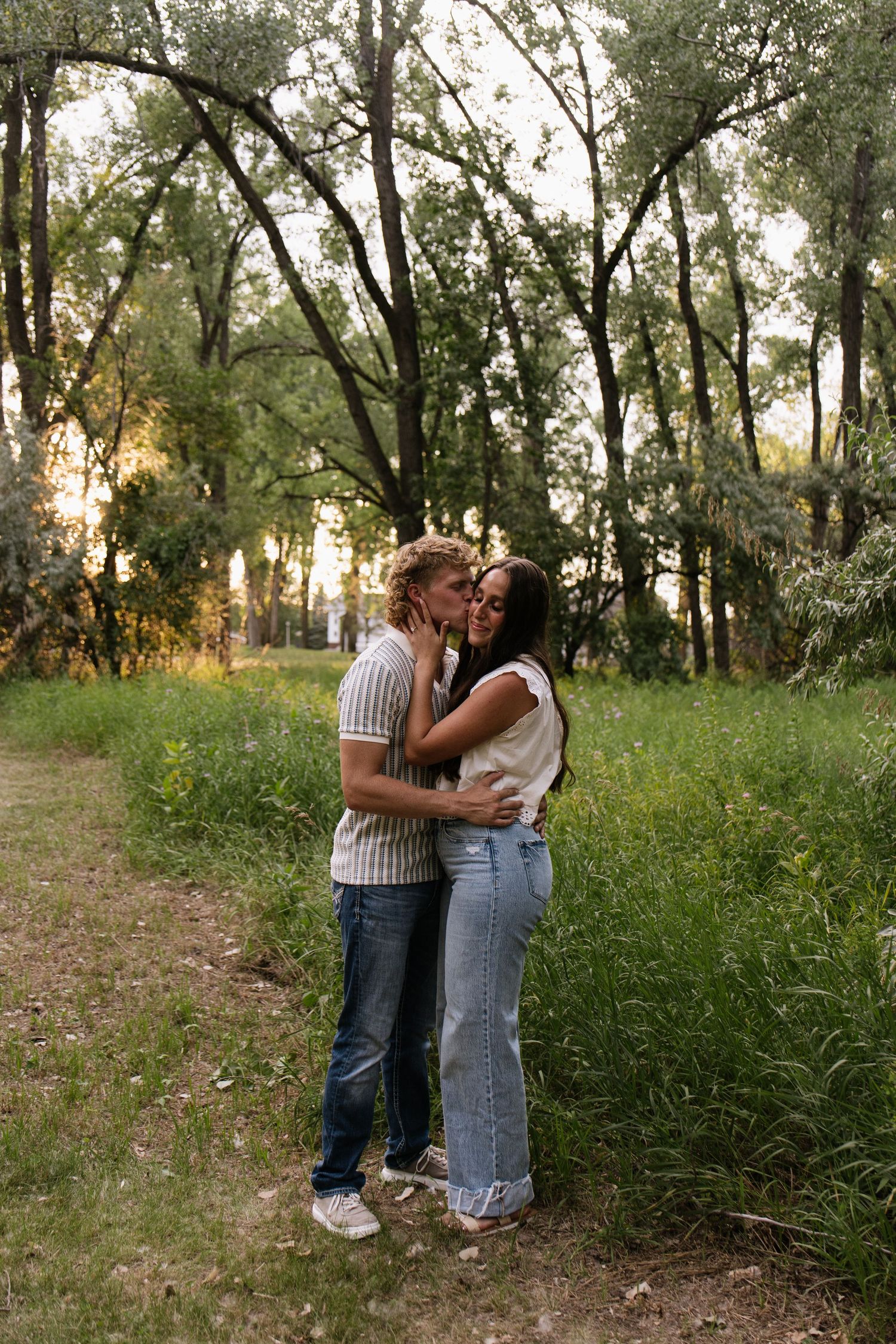 A couple embraces and kisses on a lush green park path surrounded by tall trees during golden hour.