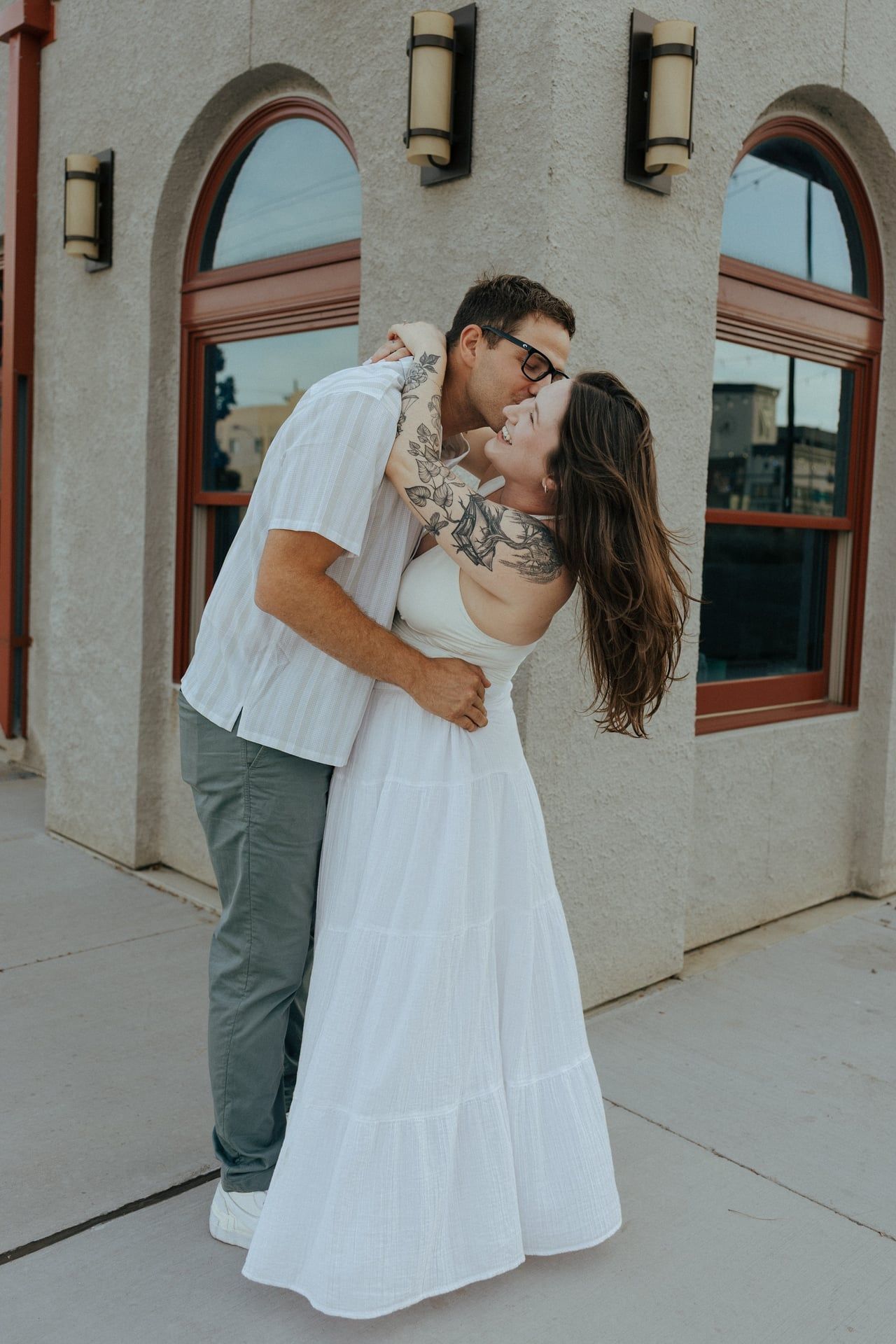 A couple shares an intimate moment against a white stucco wall with arched windows.
