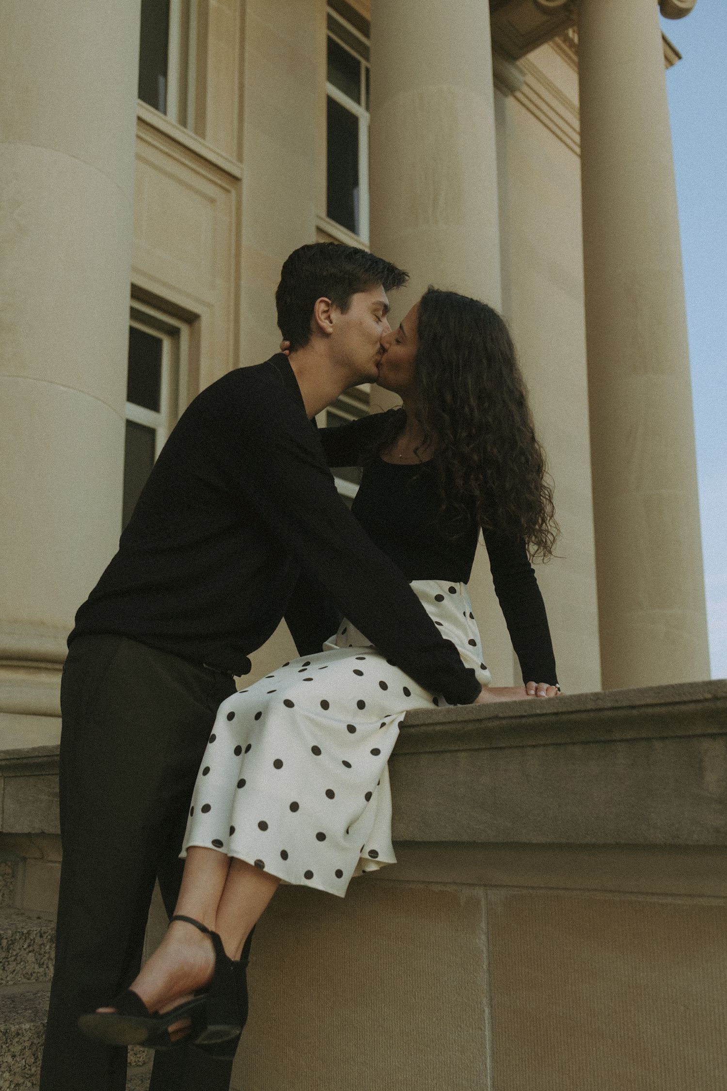 A couple shares a romantic moment against classical stone columns in formal attire.