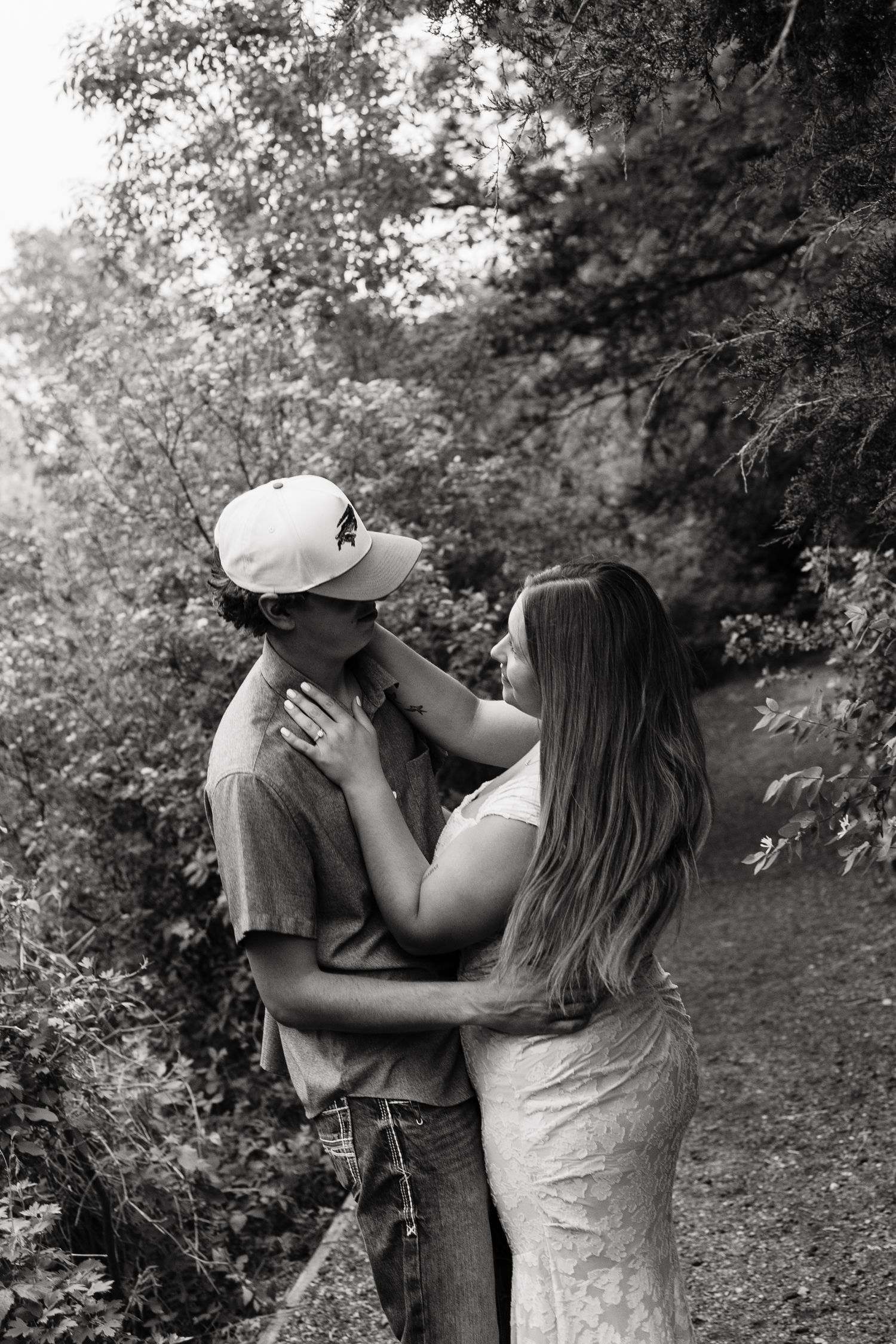 Black and white photo of a couple embracing and gazing at each other on a tree-lined path outdoors.
