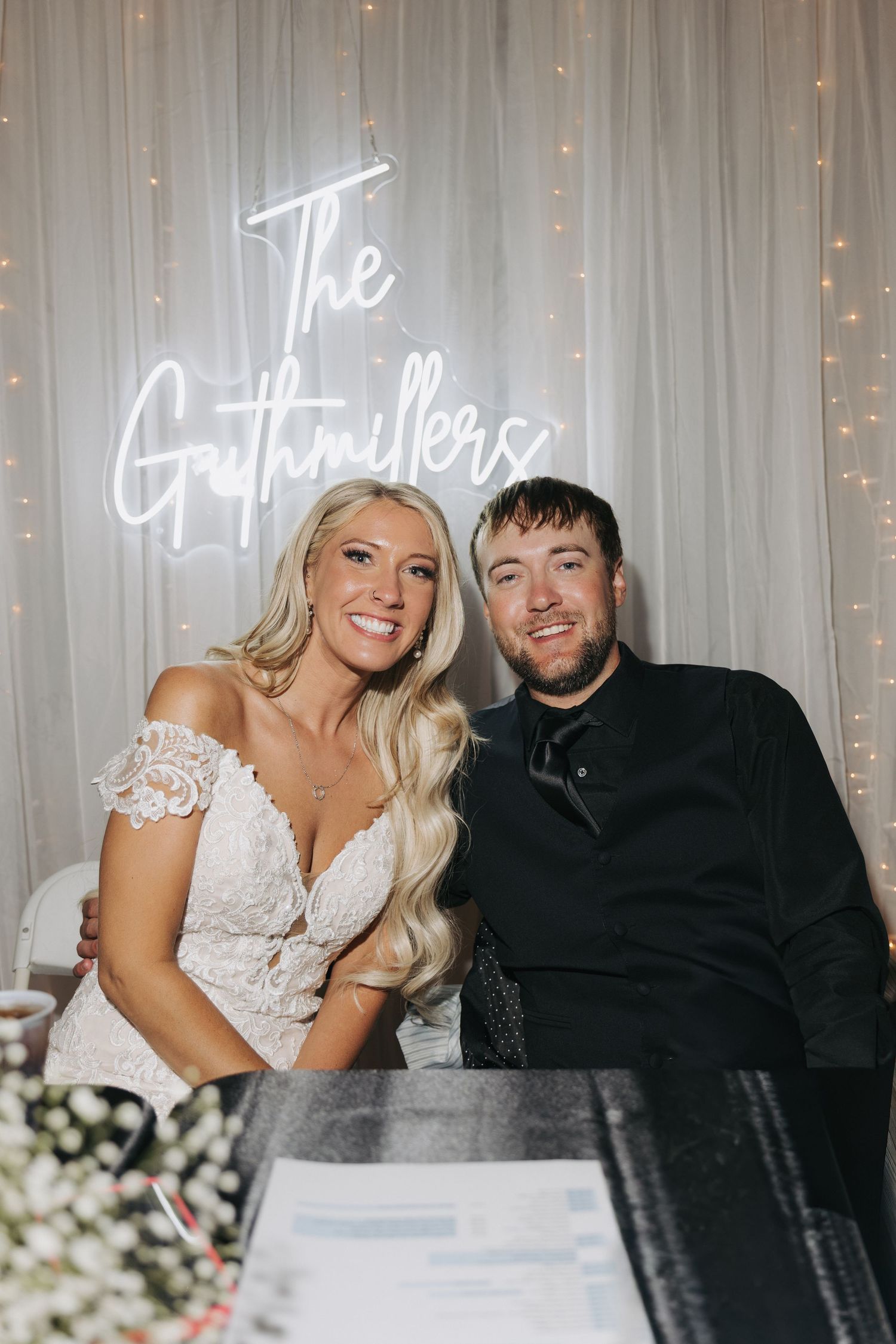 Newlyweds pose together in front of a glowing neon sign reading The Guthmillers at their wedding reception.