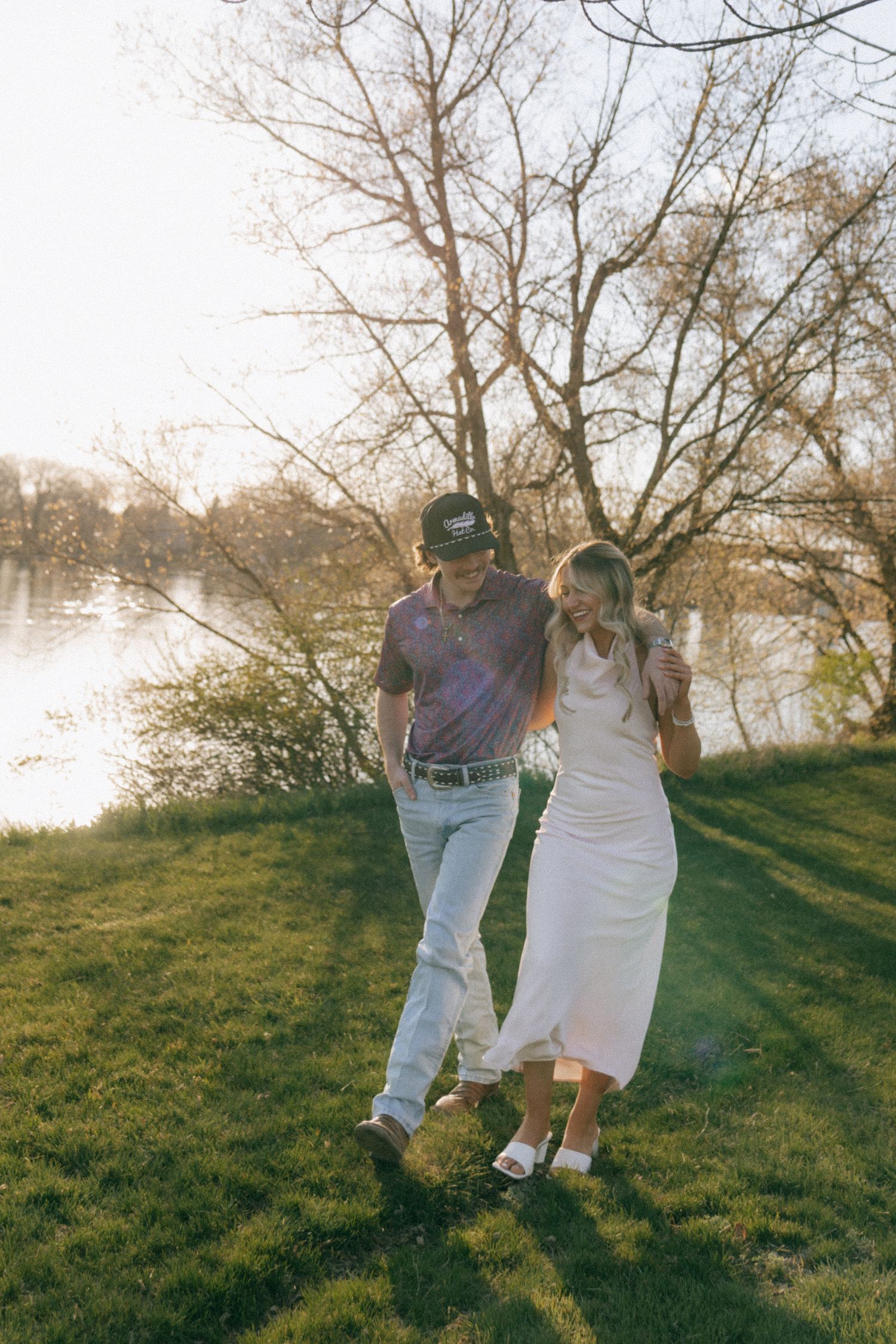Couple walking hand in hand on green grass by a river at golden hour, man in polo and cap, woman in white dress.