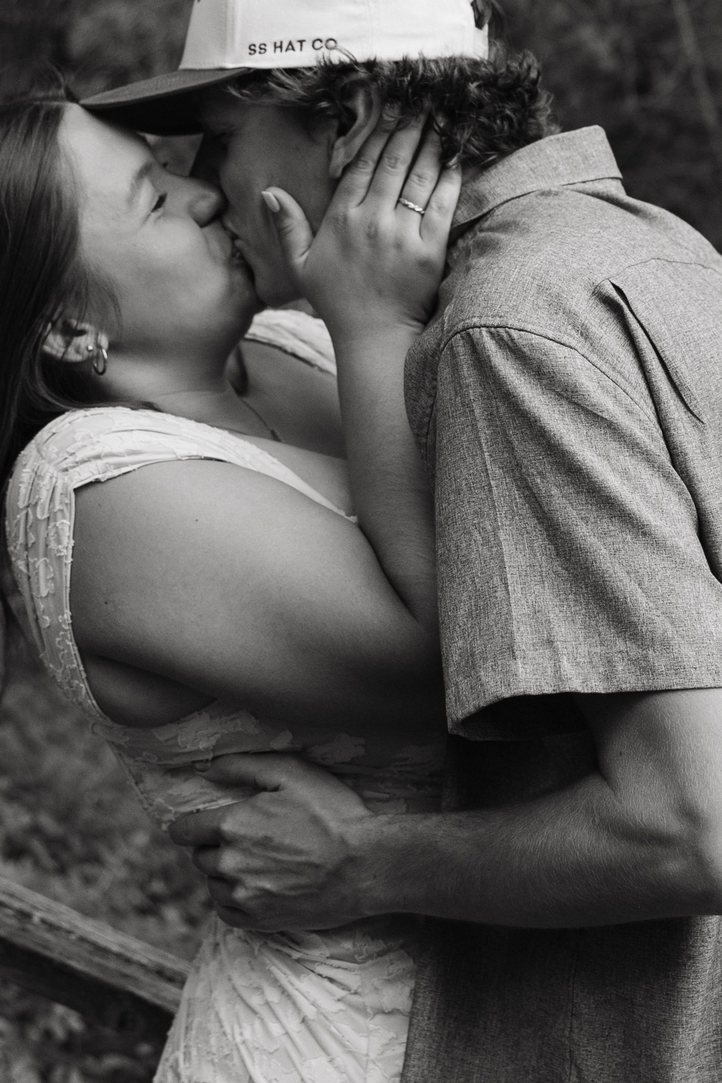 Black and white photo of a couple sharing a romantic kiss outdoors, woman in white dress, man in button-up shirt and cap.