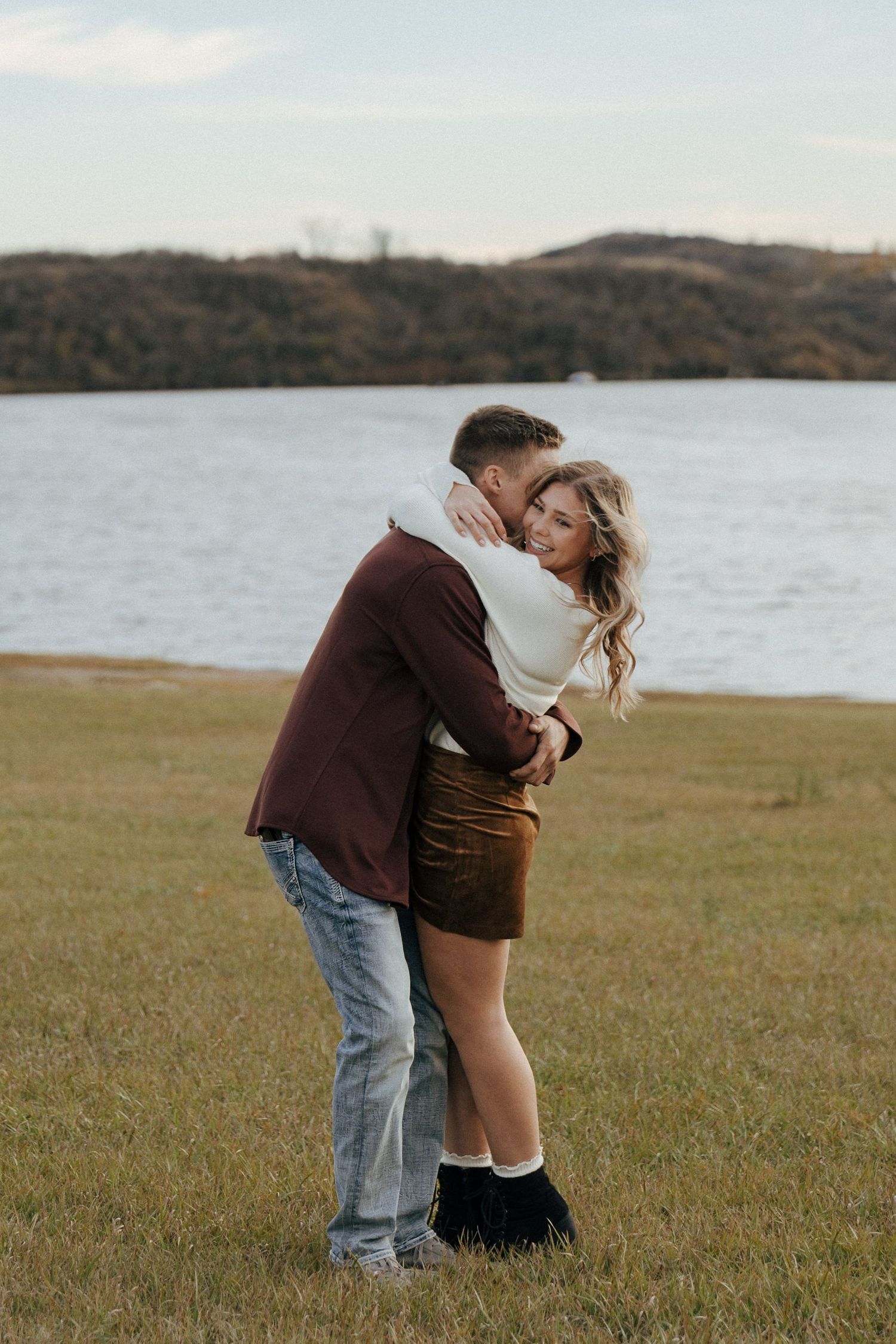 A couple embraces in a scenic field with a lake and rolling hills in the background.