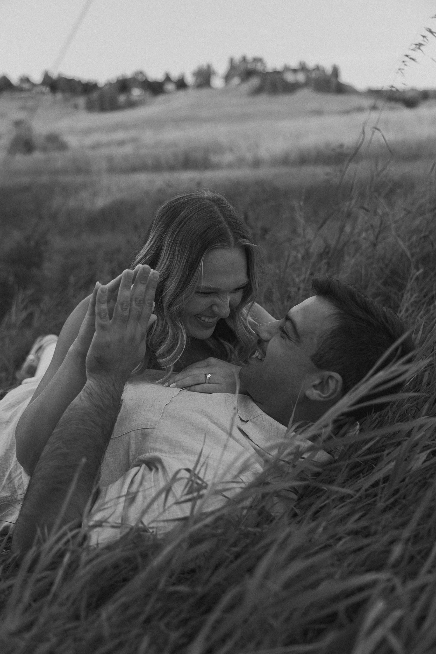 A couple shares joyful moments while lying in tall grass during an outdoor photoshoot.