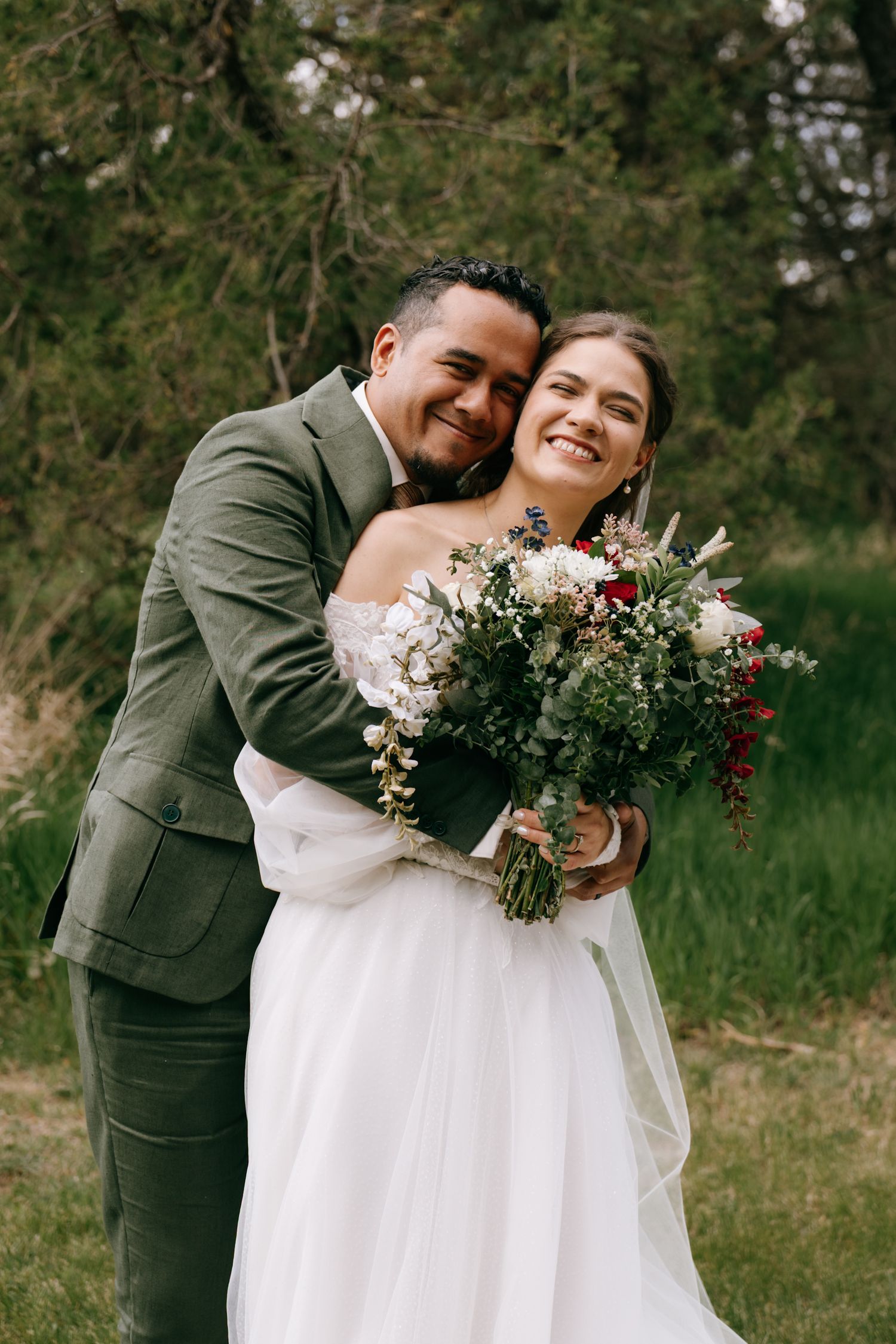 Smiling bride in white gown holds bouquet while groom in green suit embraces her outdoors.