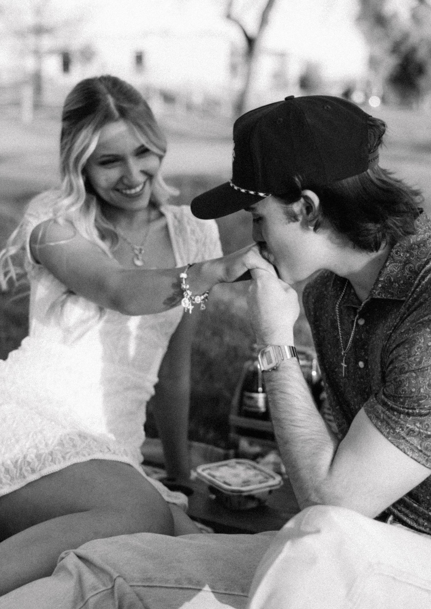 Black and white photo of a smiling blonde woman in white dress as her partner kisses her hand during a romantic picnic.