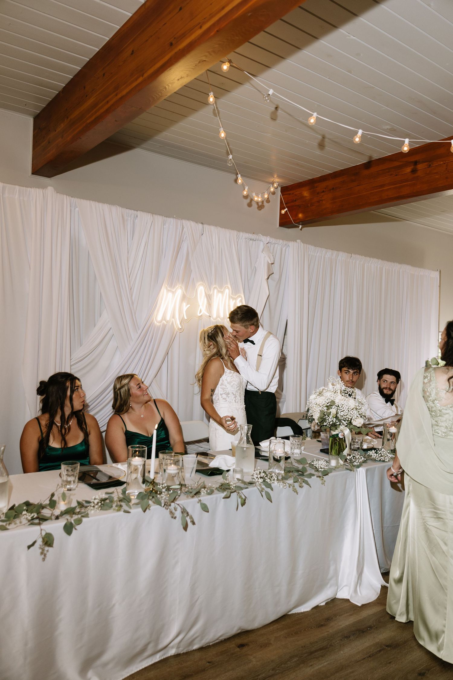 Bride and groom share a kiss at the head table during their elegant wedding reception with white draping and greenery.