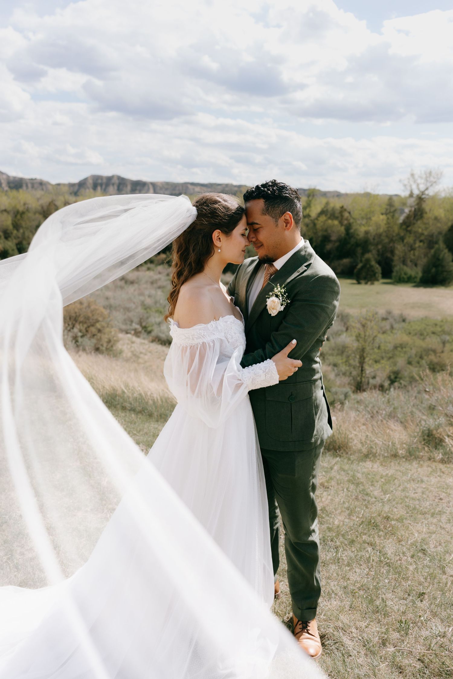 Bride and groom embrace outdoors as her veil blows in the wind, with scenic countryside views behind them.