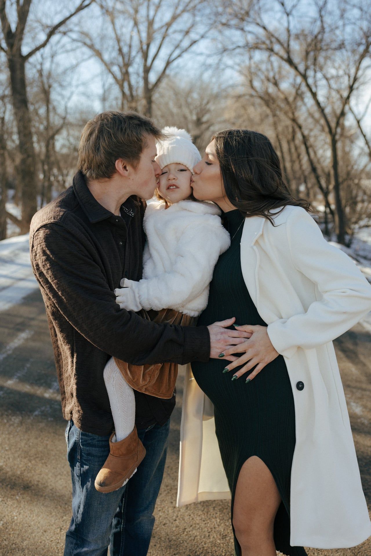 photos of mom and dad kissing cheeks of daughter in snowy trees in Bismarck