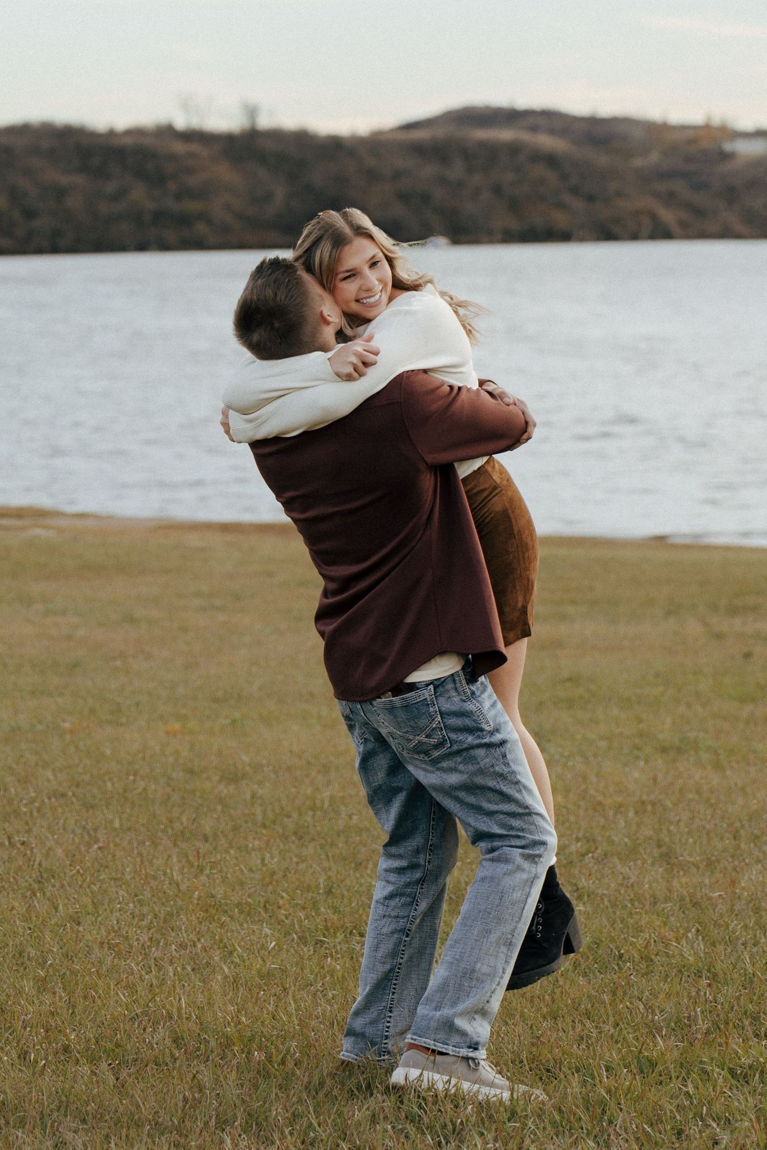 A couple shares a playful hug in a grassy field with a lake and mountains in the background.