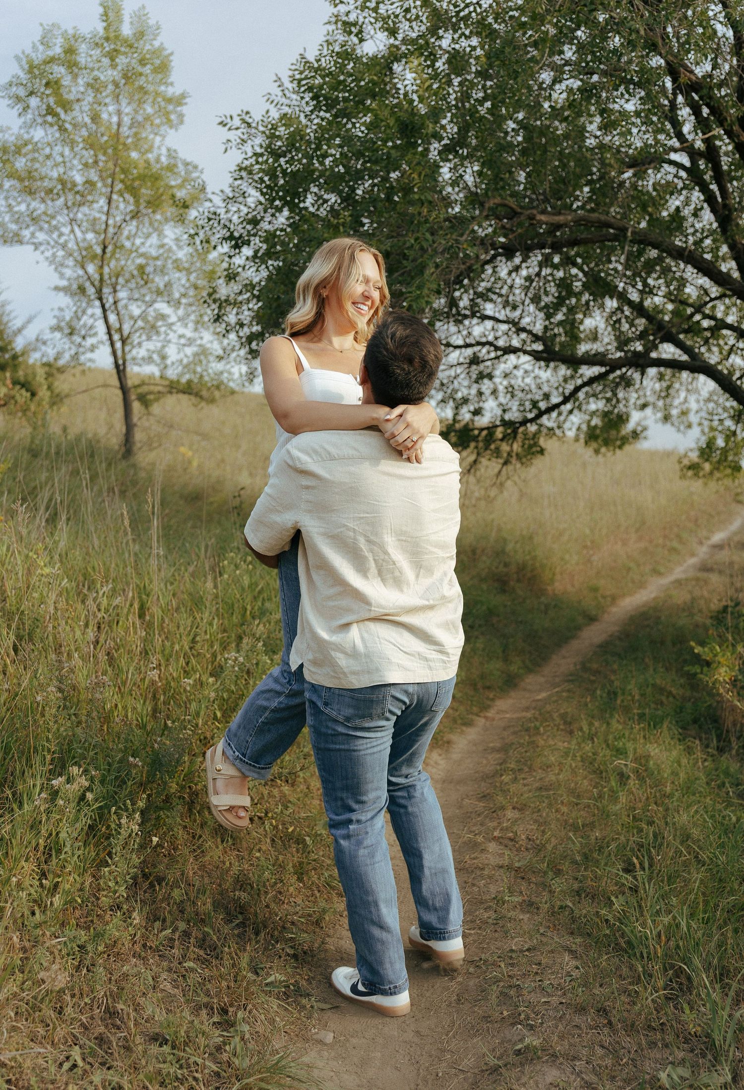 Two people walking along a dirt trail through tall grass and trees.