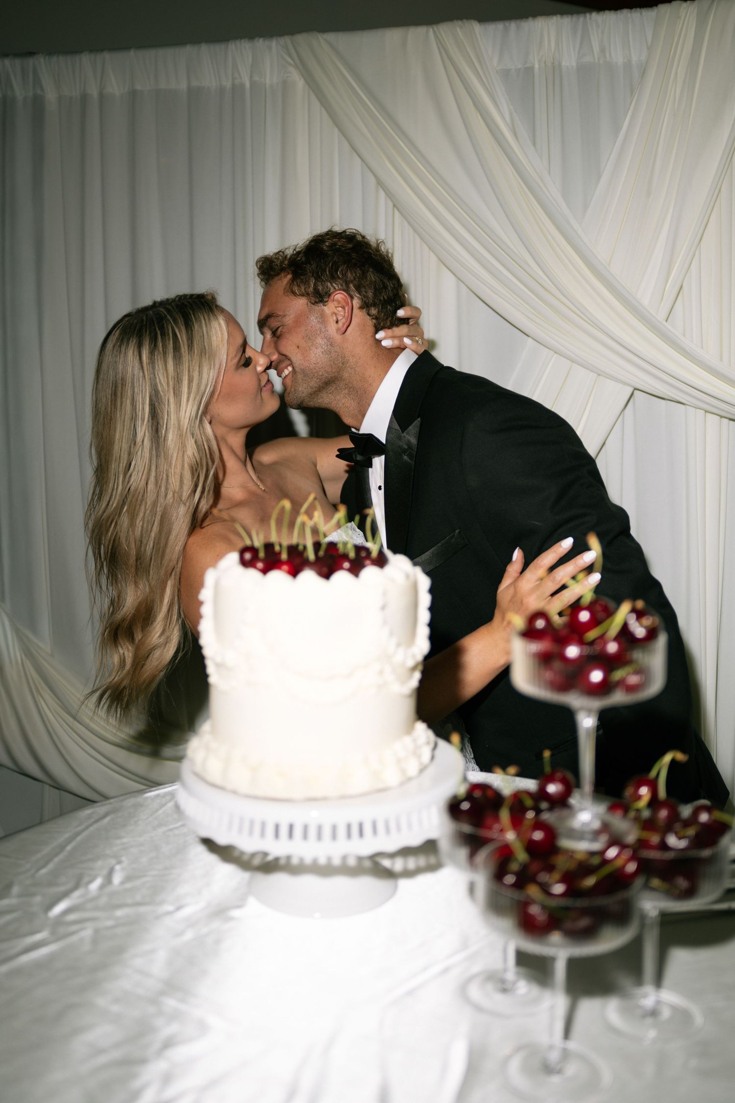 Bride and groom share a kiss behind a white wedding cake topped with cherries at their elegant reception.