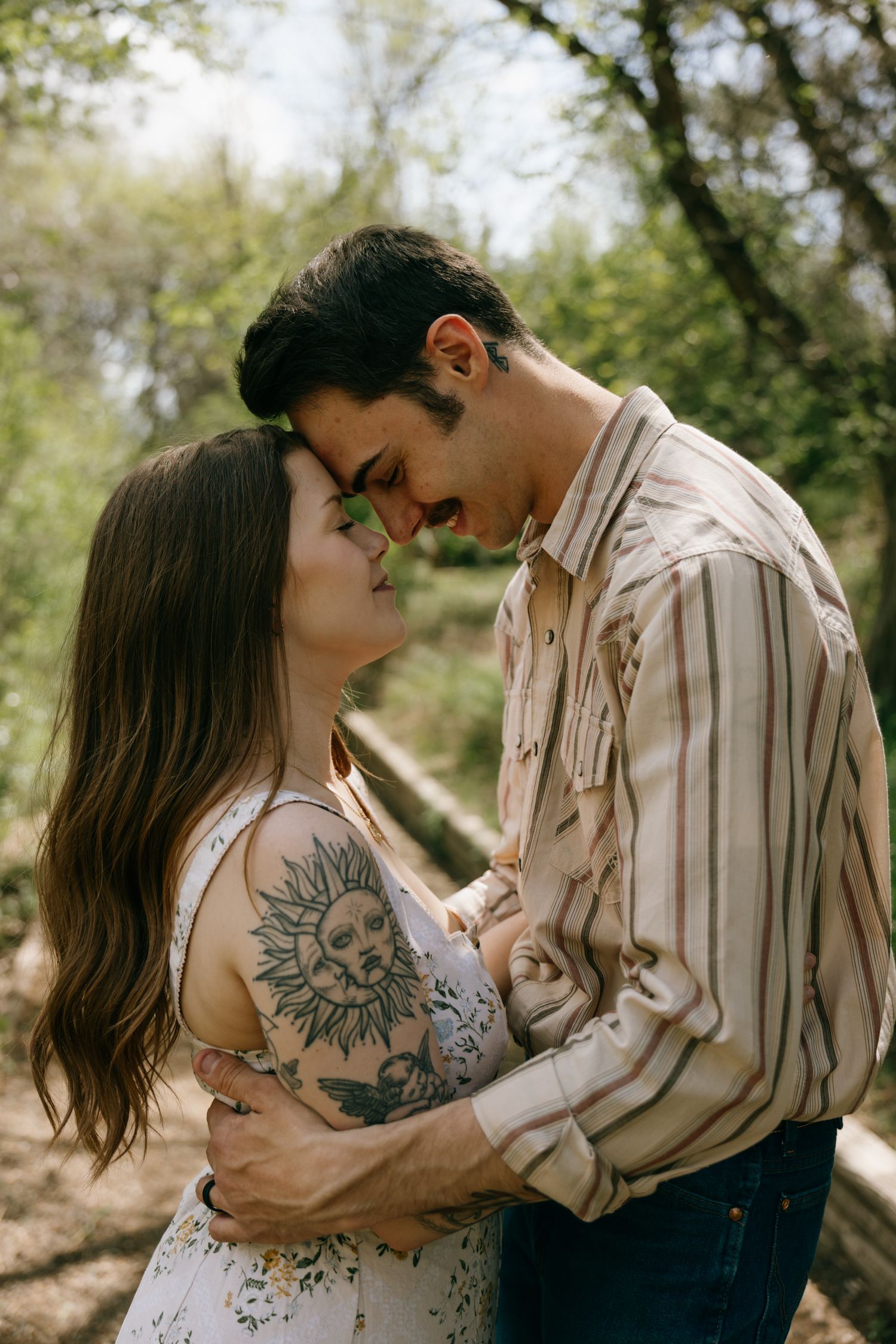 A couple with tattoos sharing a romantic moment outdoors, foreheads touching and smiling in a lush green setting.