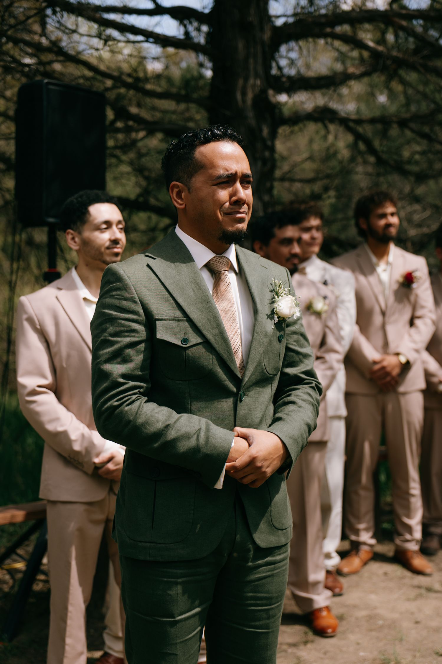Groom in green suit with boutonniere stands emotional at outdoor wedding ceremony with groomsmen in tan suits behind him.