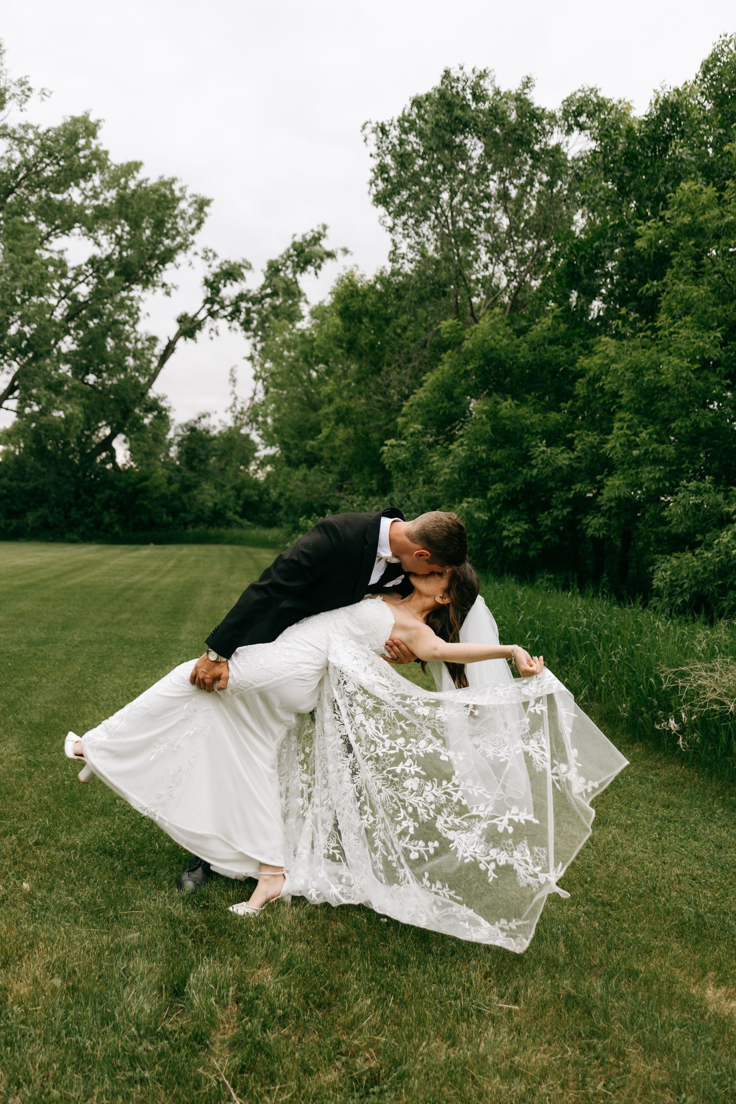 Groom dips bride for a romantic kiss on lush green grass showcasing her lace wedding gown train.