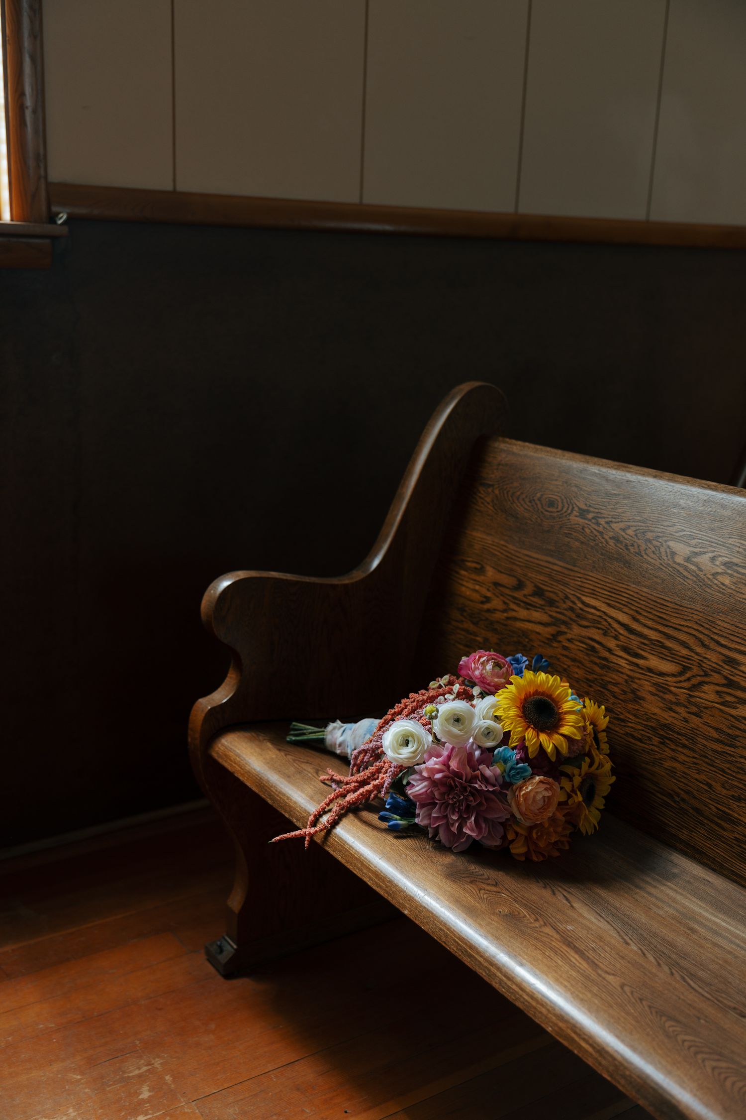 Colorful flower bouquet with sunflower resting on a wooden church pew in a dimly lit chapel.