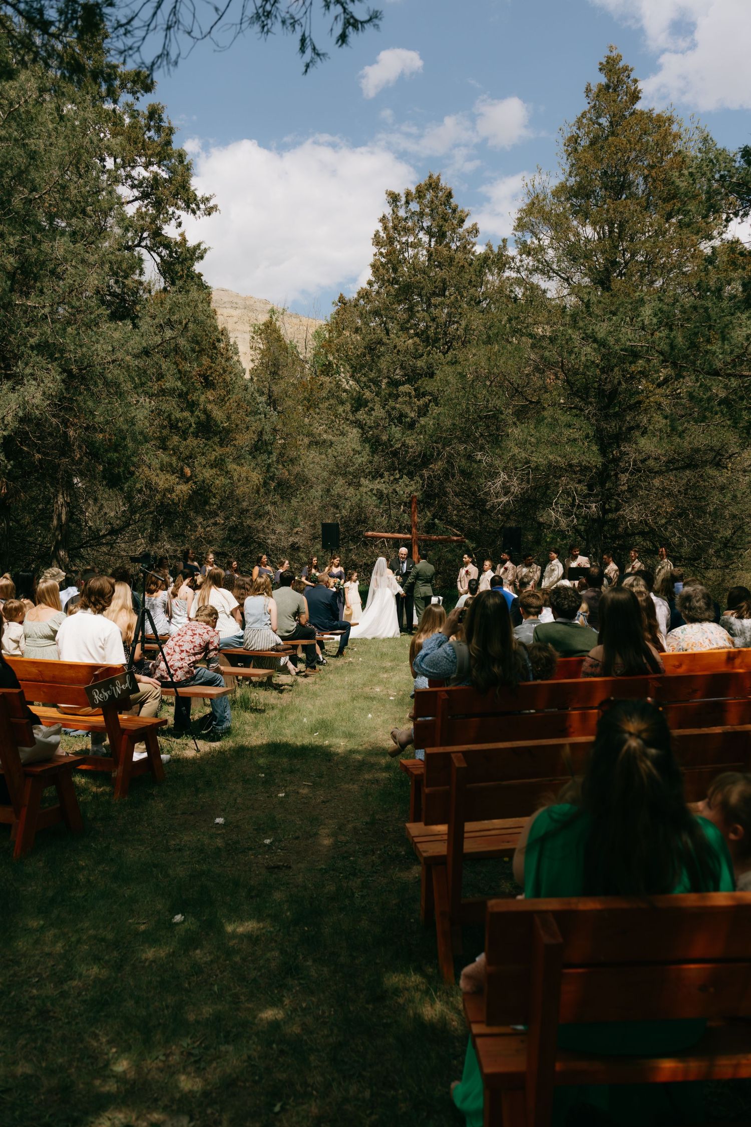 Outdoor wedding ceremony in a forest clearing with guests seated on wooden pews facing the couple at a rustic altar.
