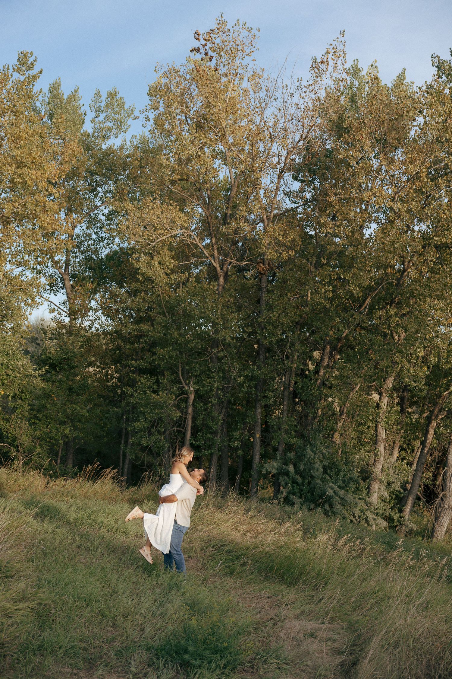 A couple playfully runs and spins through a scenic hillside with trees in the background.
