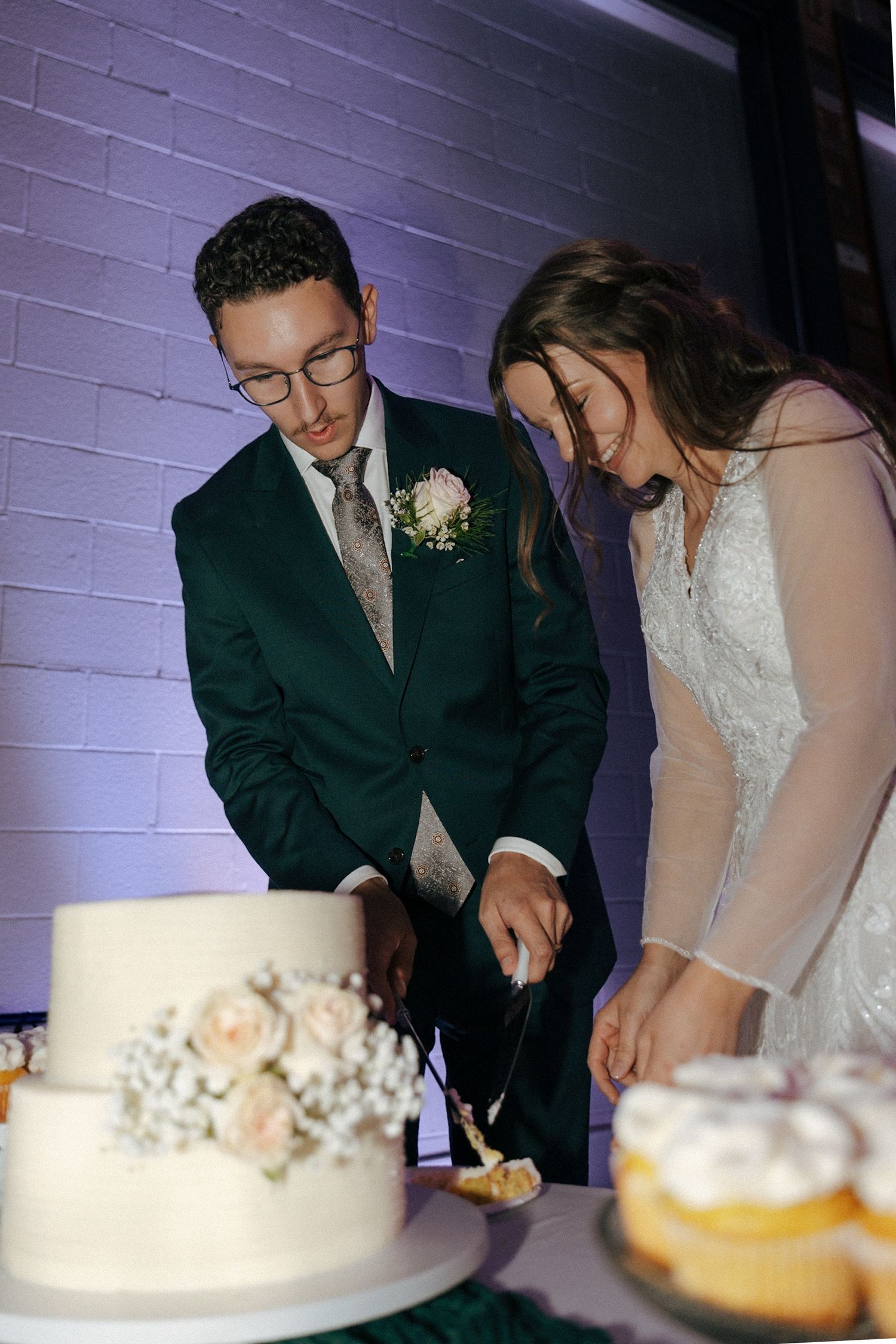 Newlyweds cut their wedding cake together surrounded by cupcakes and floral decorations.